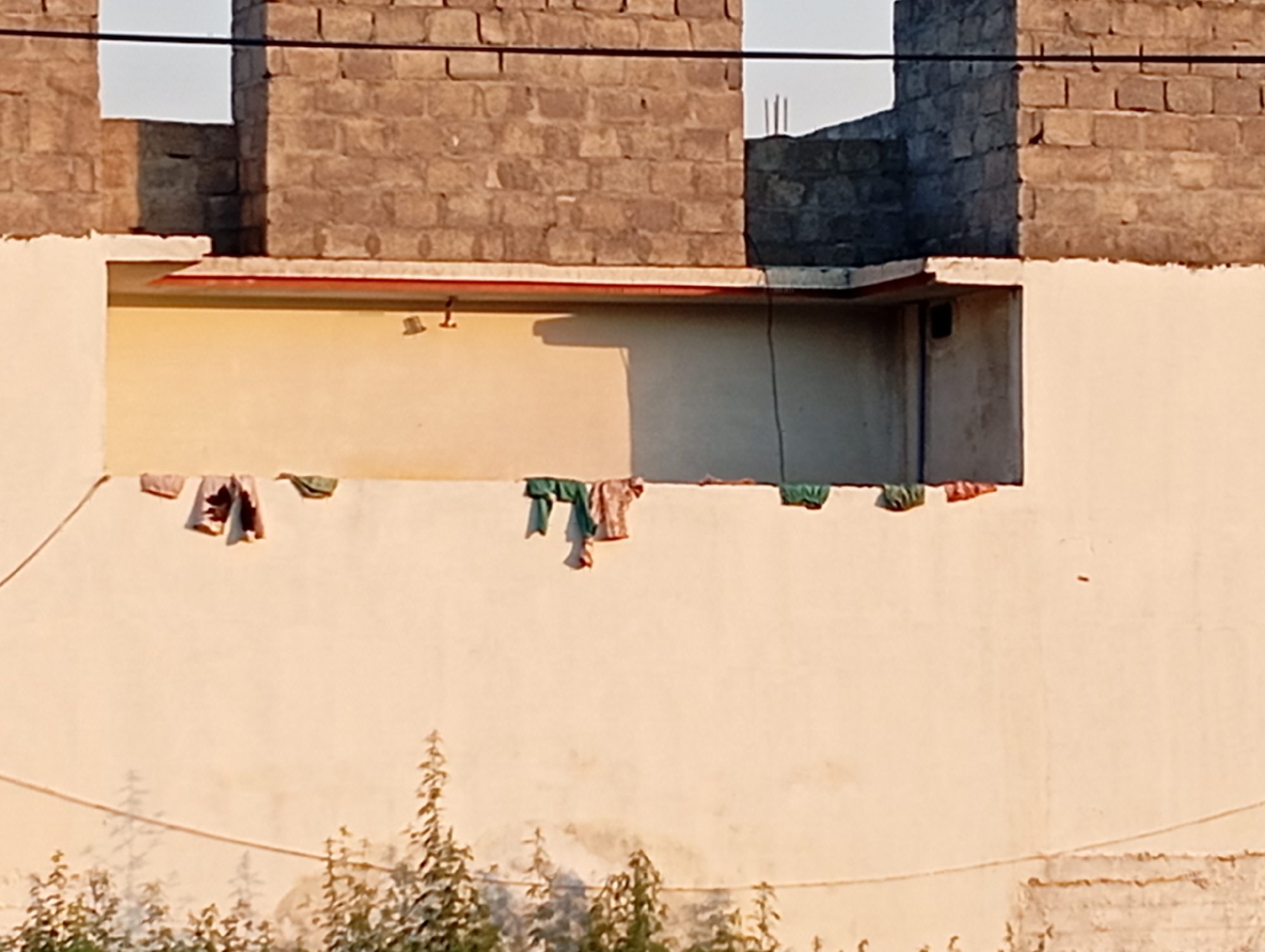 This image depicts an outdoor scene with unfinished brick structures in the background and a light-colored wall in the foreground. Several pieces of clothing are hanging on a line stretched across the wall. Below the wall, the tops of several green plants are visible, casting shadows on the wall. The overall impression is one of a residential area under construction. The image does not provide any explicit information about the city of Hakale, Pakistan, or any specific details about people, actions, or interactions.