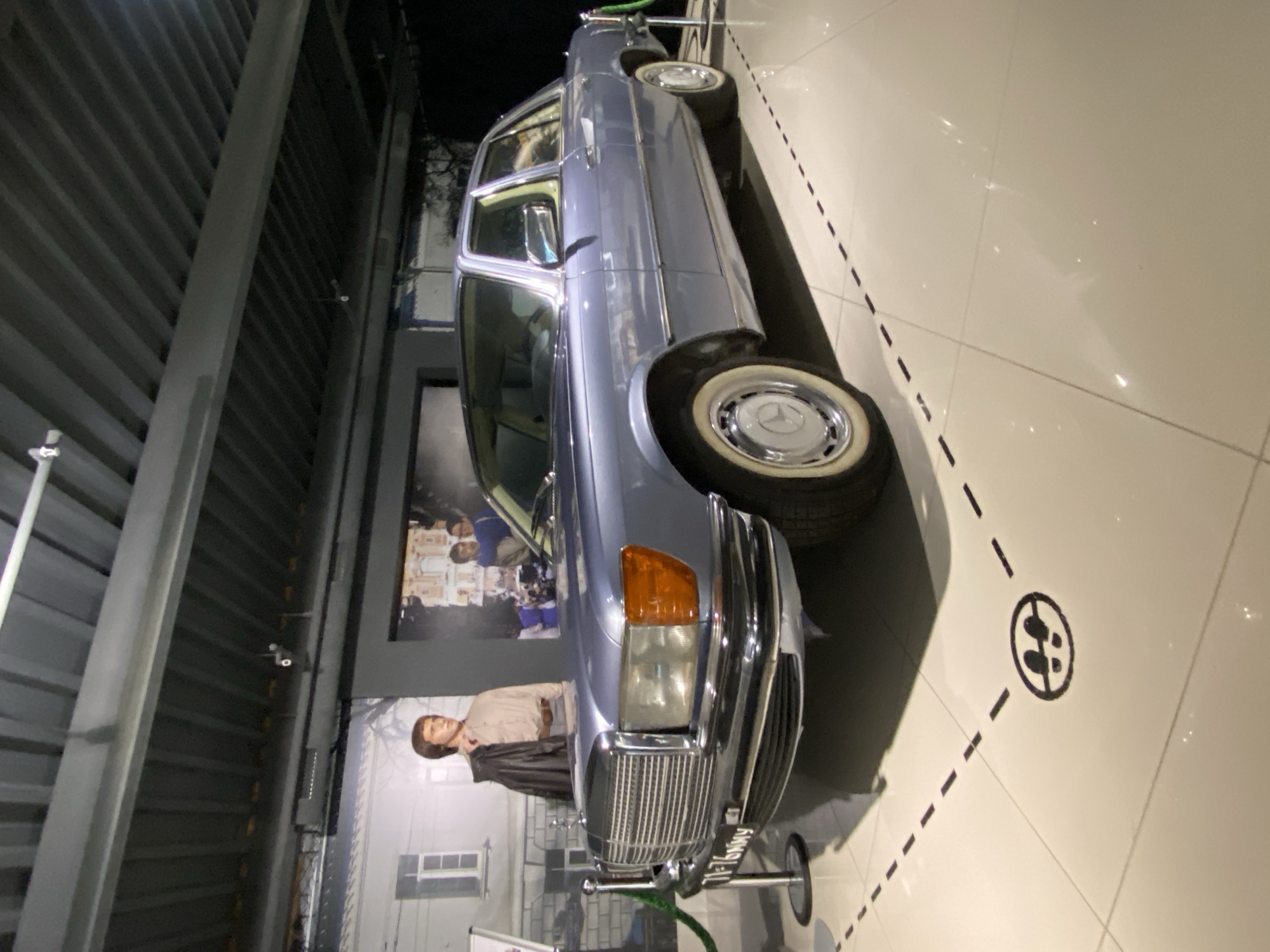 The image shows a light blue vintage Mercedes-Benz sedan displayed indoors, likely in a museum or exhibition space. The car is positioned at an angle, with its front passenger side closest to the viewer. It features a prominent chrome grille, orange turn signals, and whitewall tires. In the background, a life-sized mannequin of a man in casual 1970s attire stands beside a backdrop depicting a building and a group of people. The floor is tiled in a light color with black dashed lines forming a path and a circular symbol indicating "no walking." The overall environment suggests a controlled exhibition setting. The location of this display is the Yeltsin Center in Yekaterinburg, Russia, where the car is part of an exhibition on Boris Yeltsin's life and times.