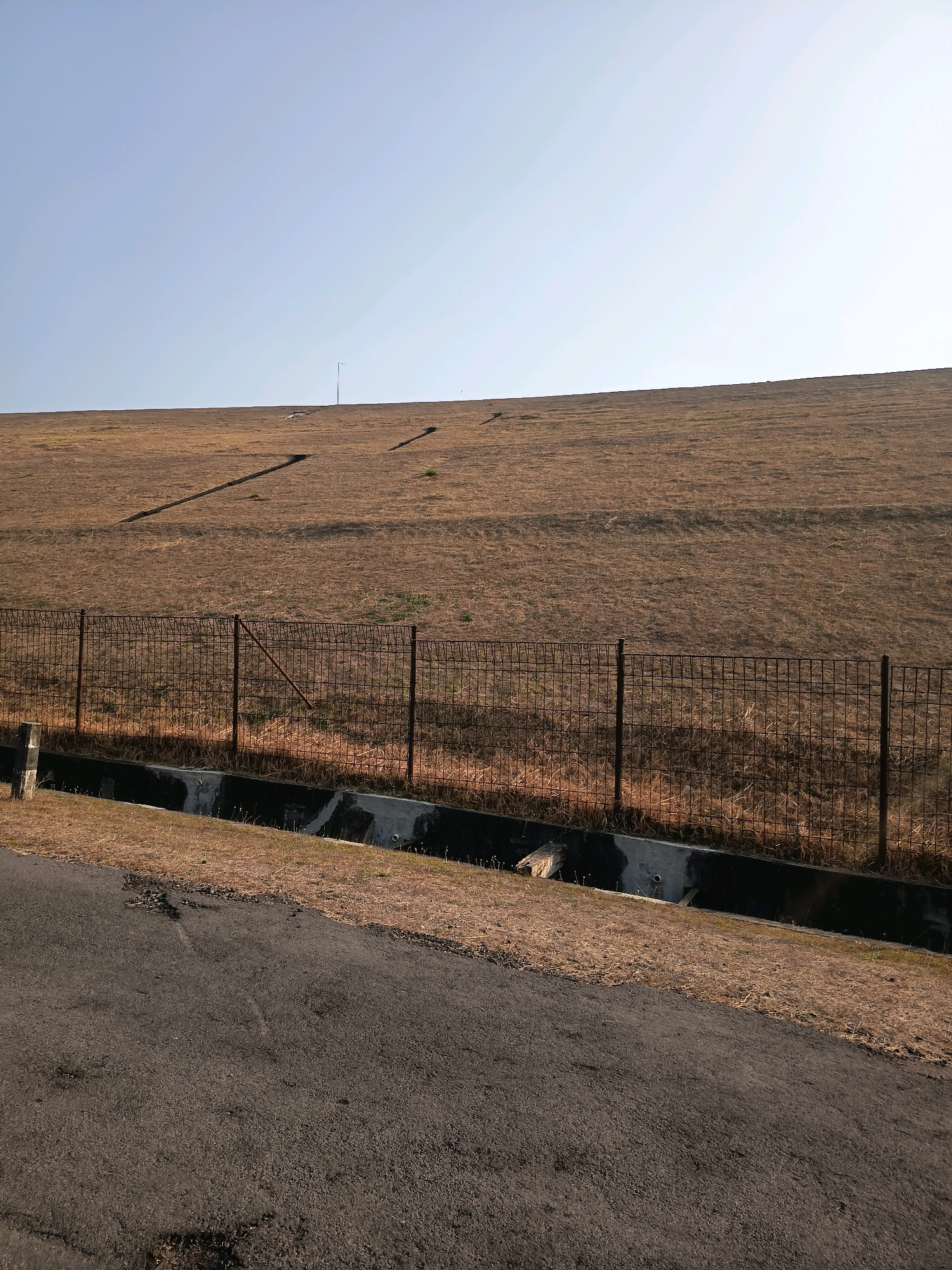 The image shows a grassy slope leading up to a clear sky. A wire fence runs horizontally across the middle ground, separating the slope from a paved area in the foreground. The slope itself is covered in dry, light brown grass, with some darker patches that appear to be bare earth or shadows. There are several diagonal lines on the slope, possibly indicating pathways or erosion control measures. In the far distance, near the top of the slope, a thin pole, possibly a lamppost, is visible. A dark-colored drainage ditch runs along the base of the slope, behind the fence. The paved area in the foreground is dark gray and appears to be a road or pathway. The overall scene is natural and uncluttered, suggesting an outdoor environment. The image is taken on a sunny day, with a bright sky.