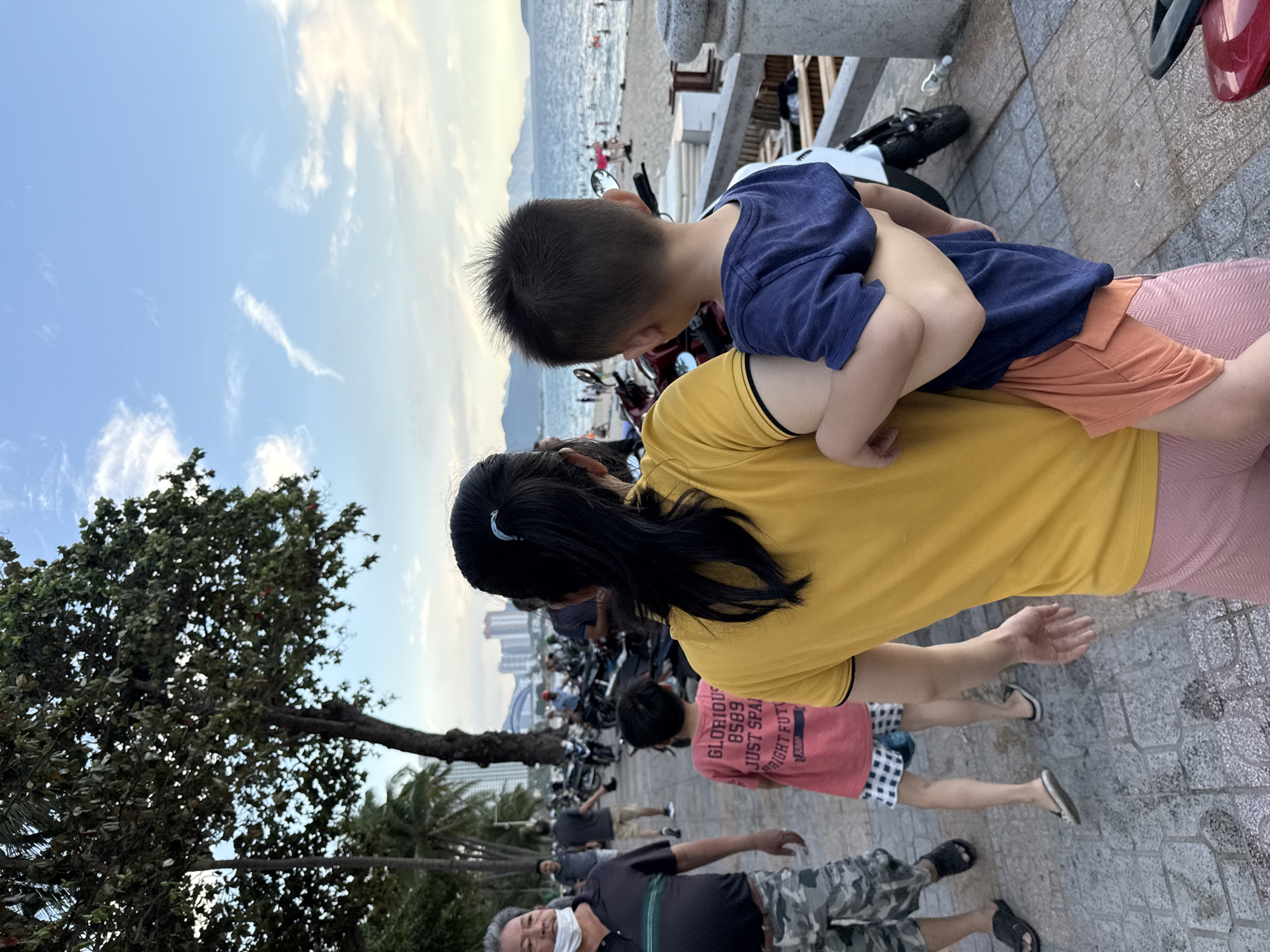 A rear view of a woman in a yellow shirt and pink shorts carrying a young child in a blue shirt and orange shorts is captured from the chest up. The child is looking to the right, out of frame. In the background, a beach with people swimming and a coastline with buildings and mountains are visible under a blue sky with scattered clouds. A man in a black shirt and camouflage shorts, wearing a mask around his neck, walks to the left. A child in a pink t-shirt and patterned shorts walks in front of the woman and child. Several motorcycles are parked on the right side of the frame. The scene is set on a paved walkway in Nha Trang Ward, Vietnam, near a beach.