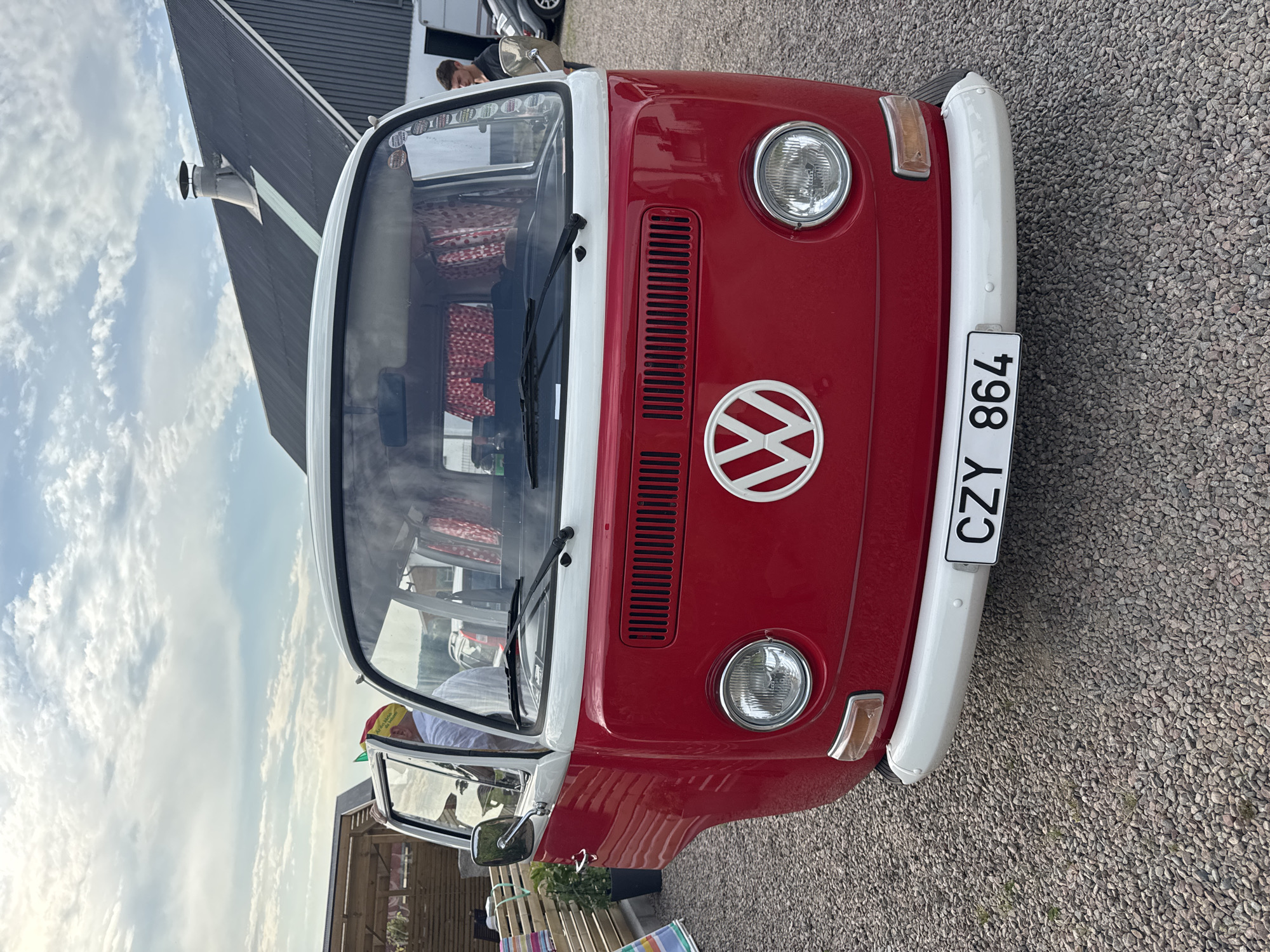 This media file shows a close-up, head-on view of a red and white Volkswagen Type 2 (Kombi) parked outdoors on a gravel surface. The van's front is the primary focus, with its distinctive round headlights, turn signals, and the iconic VW logo visible. The license plate reads "CZY 864". Inside the van, through the windshield, red patterned curtains are visible, suggesting it might be used as a camper or has a custom interior.
In the background, a modern building with a dark, corrugated metal roof is partially visible, as is a section of a trailer. A person is leaning out of the driver's side window, wearing a white shirt and a colorful hat. Another person is partially visible in the background, standing near the trailer. The sky is cloudy with some patches of blue, indicating daytime. The lighting suggests it could be late afternoon or a bright, overcast day.
The overall scene appears to be an outdoor event or gathering, possibly a car show or a community event, given the presence of multiple vehicles and people. The setting in Vessigebro, Sweden, suggests a Northern European environment. There is a sense of casual interaction and enjoyment associated with vintage vehicles. Visible text includes the license plate "CZY 864" and stickers on the top of the windshield.
