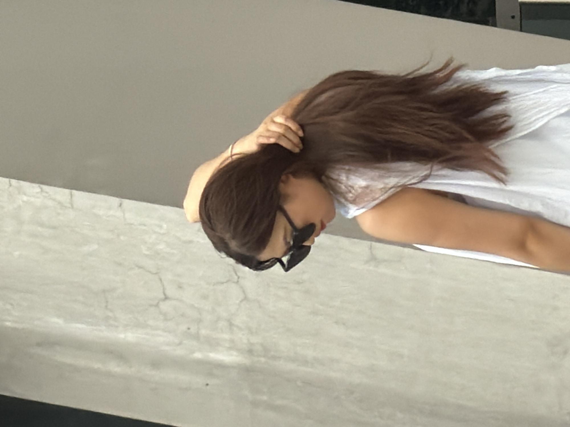 A medium-shot, profile view shows a woman from the shoulders up, facing right, with her head tilted down and her right hand running through her long, brown hair. She is wearing dark, angular sunglasses and a white top with lace details. The background consists of a textured, light-colored concrete wall on the left and a plain, grey surface on the right. The scene appears to be outdoors. No Yuzhong District, China is specified in the image.