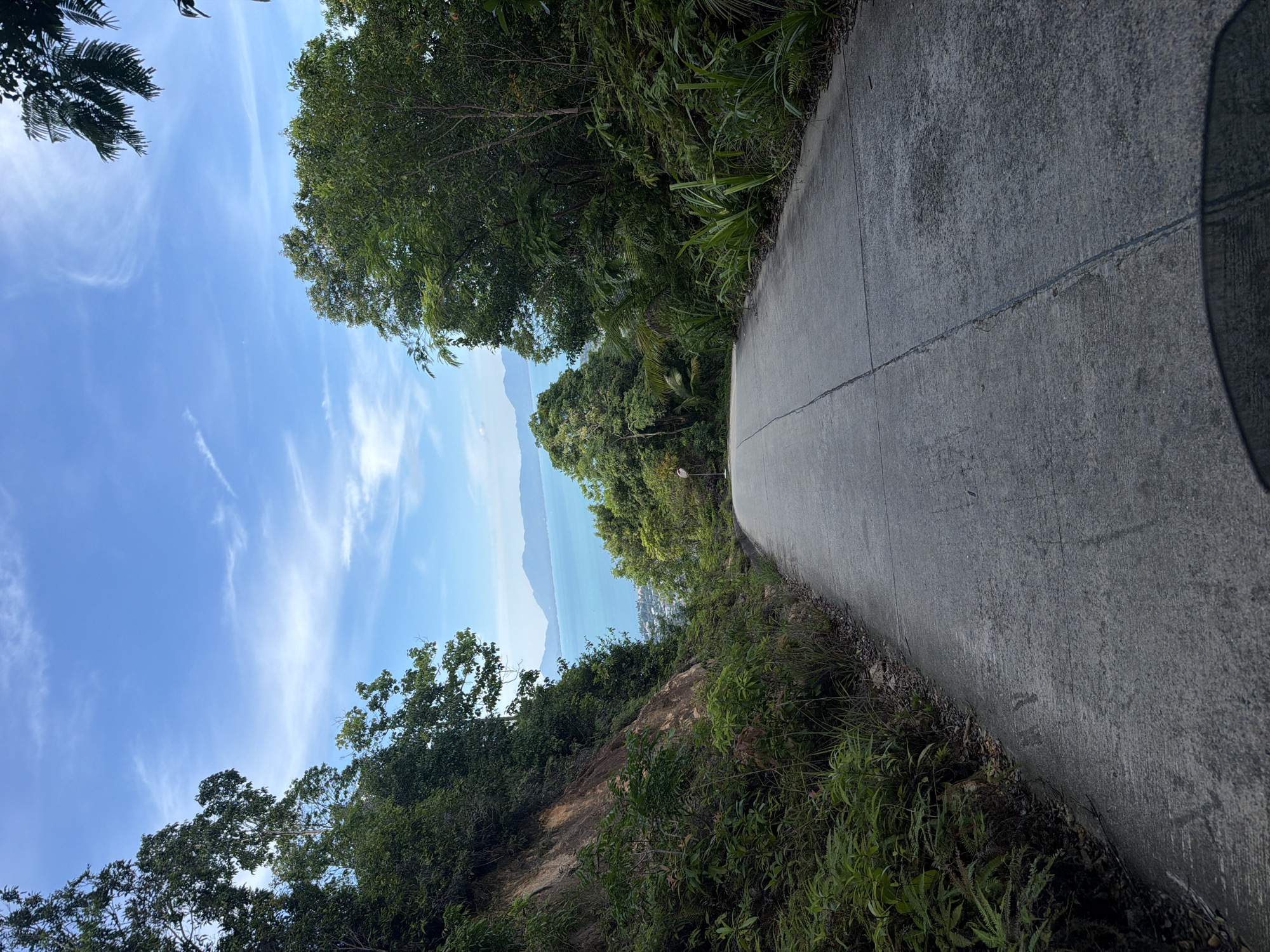 A paved, two-lane road slopes gently uphill and curves to the right, surrounded by dense green foliage. The road is made of concrete with a visible seam running down the center. To the left, a rocky, vegetated hillside rises sharply. To the right, more trees and plants line the roadside. In the distance, beyond a break in the trees, a bright turquoise ocean is visible, with islands or mountains on the horizon under a blue sky with scattered white clouds. The scene appears to be a scenic overlook or a road leading to one in a tropical environment. No people or specific man-made structures, other than the road, are clearly identifiable. The overall impression is one of a natural, elevated landscape with a view of the sea.