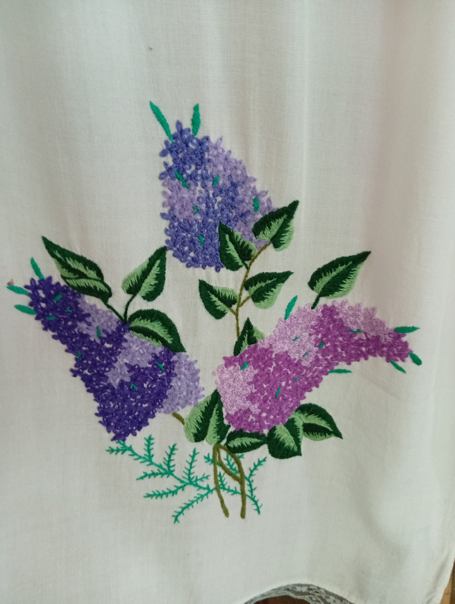 The image shows an embroidered pattern of three lilac branches on a white fabric. The branches are depicted with varying shades of purple and green embroidery. The fabric appears to be a natural material, possibly linen or cotton, and is slightly creased, suggesting it might be a piece of clothing or home decor. The background is plain white. There is no information about the location in the city of Крыўлянскі сельскі Савет, Belarus.