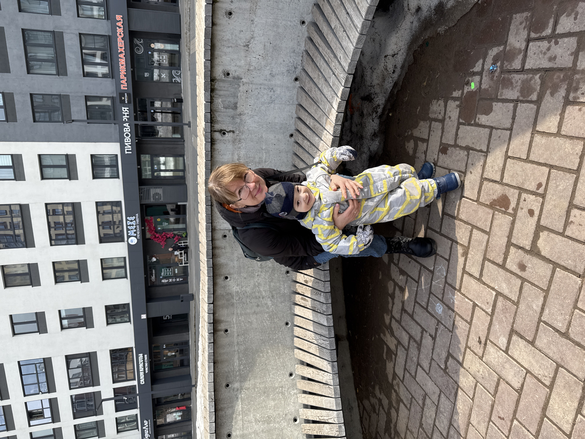 The image captures an adult female and a young child standing in a paved urban area on K. Marx Street, 230, Izhevsk, Russia.
The adult female, positioned on the left, wears a dark jacket, blue jeans, and black boots. She has light brown hair, wears eyeglasses, and a dark backpack is visible on her left shoulder. She is smiling and looking directly at the camera, with her right arm supporting the child at the waist.
The child, a toddler, is positioned in front of the adult, wearing a grey and yellow patterned full-body snowsuit, a blue and grey knitted hat, and blue boots. The child is also smiling and looking directly at the camera, standing in an upright posture.
Behind the individuals is a curved concrete structure featuring an integrated wooden slatted bench. The concrete exhibits a raw finish with circular formwork marks.
The ground consists of light-colored brick paving stones arranged in a herringbone pattern. Visible on the ground are several wet patches, scattered dirt, small debris (including a green object, a blue object, and white chalk markings).
In the background, a multi-story modern building features alternating white and dark grey sections with numerous rectangular windows. The ground floor comprises various commercial establishments with dark fascias. Identifiable signs include "БУНГАЛО," "САЛОН КРАСОТЫ ЧУВСТВО СТИЛЯ," "MEZE ГРЕЧЕСКАЯ ТАВЕРНА" (adorned with a red floral arrangement), "ПИВОВА РНЯ," and "ПАРИКМАХЕРСКАЯ." The scene is illuminated by natural daylight.