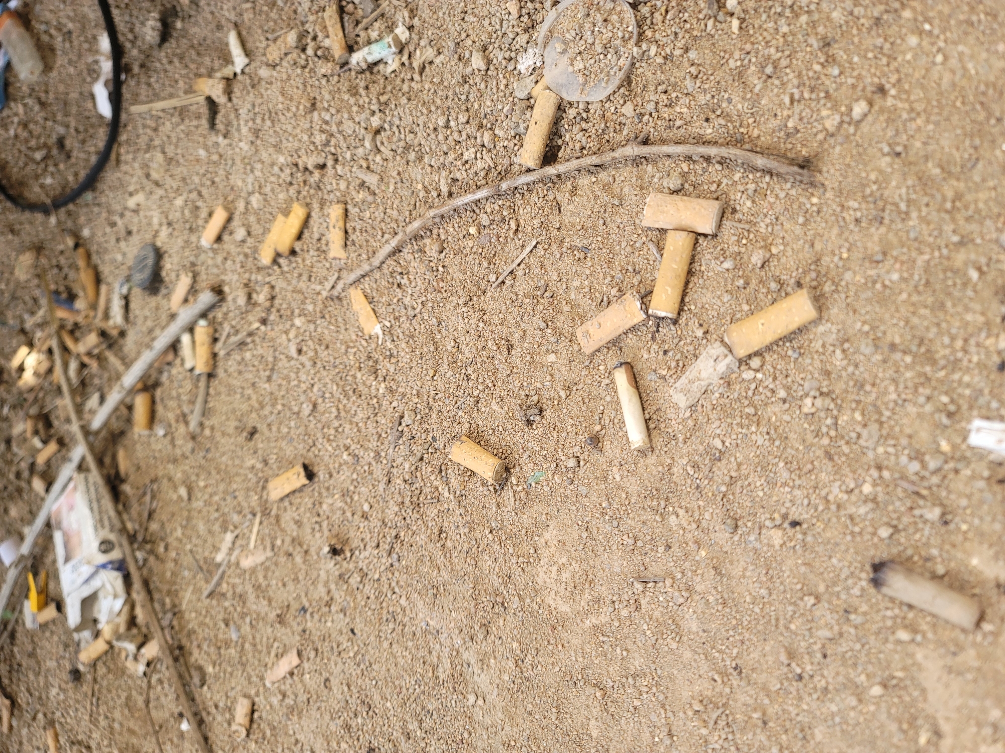 The image depicts a close-up, overhead view of a patch of dry, sandy ground littered with numerous cigarette butts, debris, and a broken pack of cigarettes. The ground is covered in small pebbles and dirt. Among the cigarette butts are a few small twigs and what appears to be a plastic cap from a bottle or a disposable lighter. In the upper left of the image, a partially obscured cigarette pack is visible, along with some crumpled paper and a yellow plastic object. The overall impression is one of neglect and pollution. This scene could be found in an outdoor public space in Jos, Nigeria, such as a street corner, a park, or an unkempt area. No individuals are present in the image. The lighting suggests it is daytime.