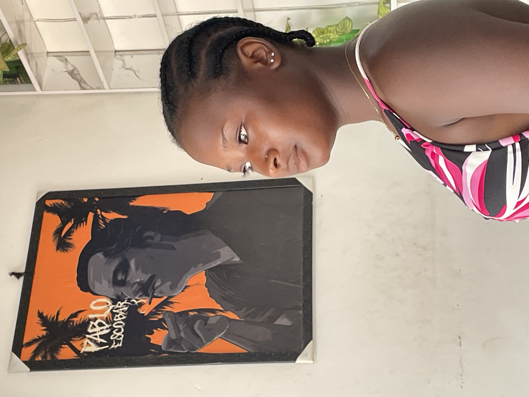 The media file displays an indoor scene, presented rotated 90 degrees clockwise. In its intended upright orientation, a young Black person, likely female, is captured from the chest up, looking thoughtfully downwards. She has dark skin, hair styled in cornrow braids, and wears two silver stud earrings in her left earlobe along with a small silver nose stud. A delicate gold chain necklace is visible around her neck, and she is dressed in a vibrant top featuring a pattern of black, pink, and white abstract shapes.
Behind and to the left of the person, a framed painting hangs on a light-colored wall. The artwork is a stylized pop-art depiction of a man, rendered in shades of gray and black, against a bright orange background. Silhouettes of palm trees frame the figure, and the name "PABLO ESCOBAR" is written in bold white script below the man's chin. The upper background reveals a white ceiling with linear paneling and glimpses of green plants, suggesting a bright, daytime setting in Barakin Akawo, Nigeria. The overall ambiance is casual and personal.