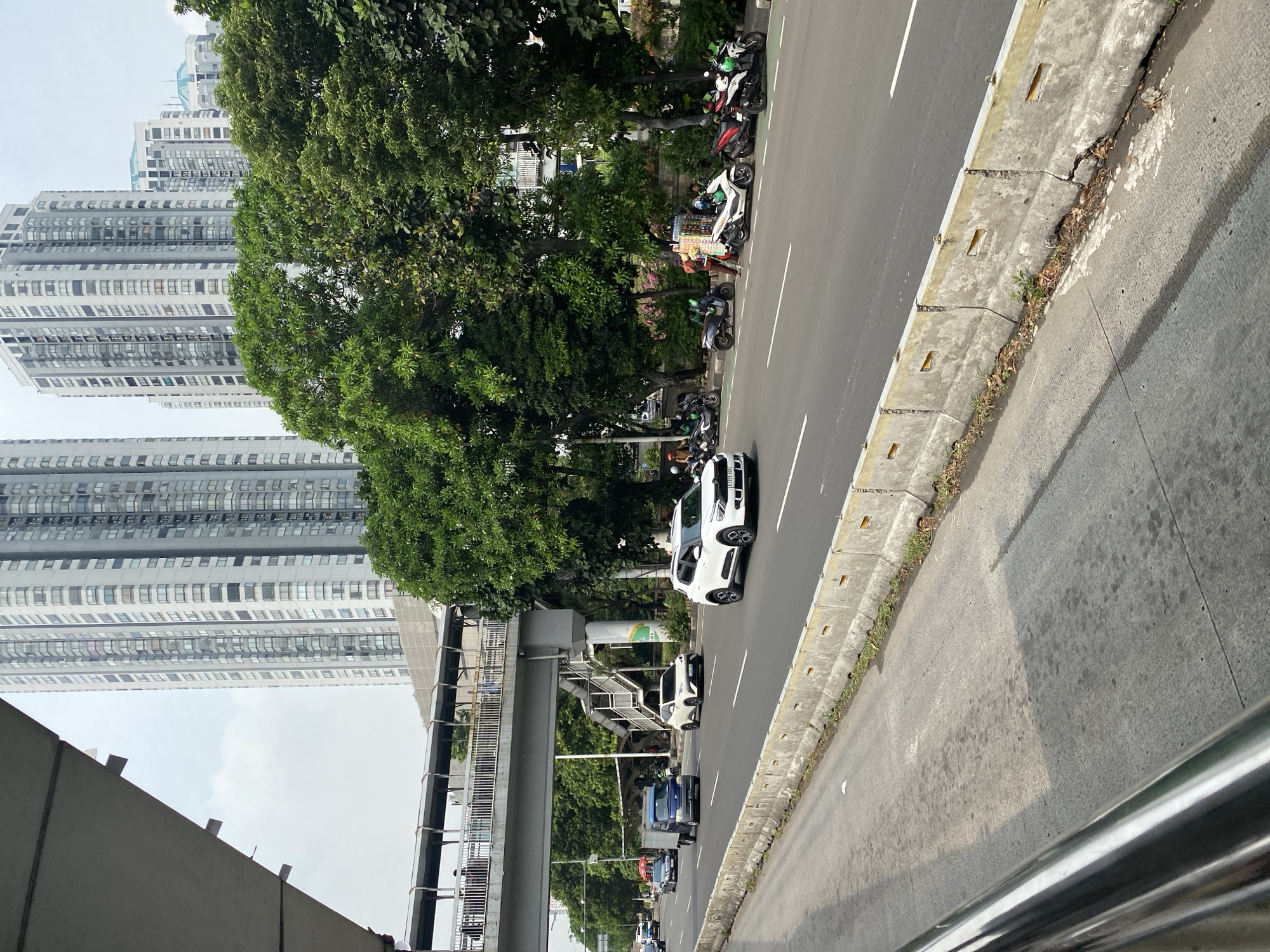 In West Jakarta, Indonesia, a wide, multi-lane road is depicted, separated by concrete barriers. Several vehicles, including cars and motorcycles, are in motion on the road. A pedestrian bridge arches over the street in the middle ground, with several people visible walking across it. Tall, modern buildings with many windows rise in the background, characteristic of a dense urban environment. Lush green trees line the side of the road, providing a contrast to the concrete structures. Along the side of the road, several people are gathered near parked motorcycles, some appearing to be vendors displaying goods. The sky is overcast, suggesting a cloudy day.