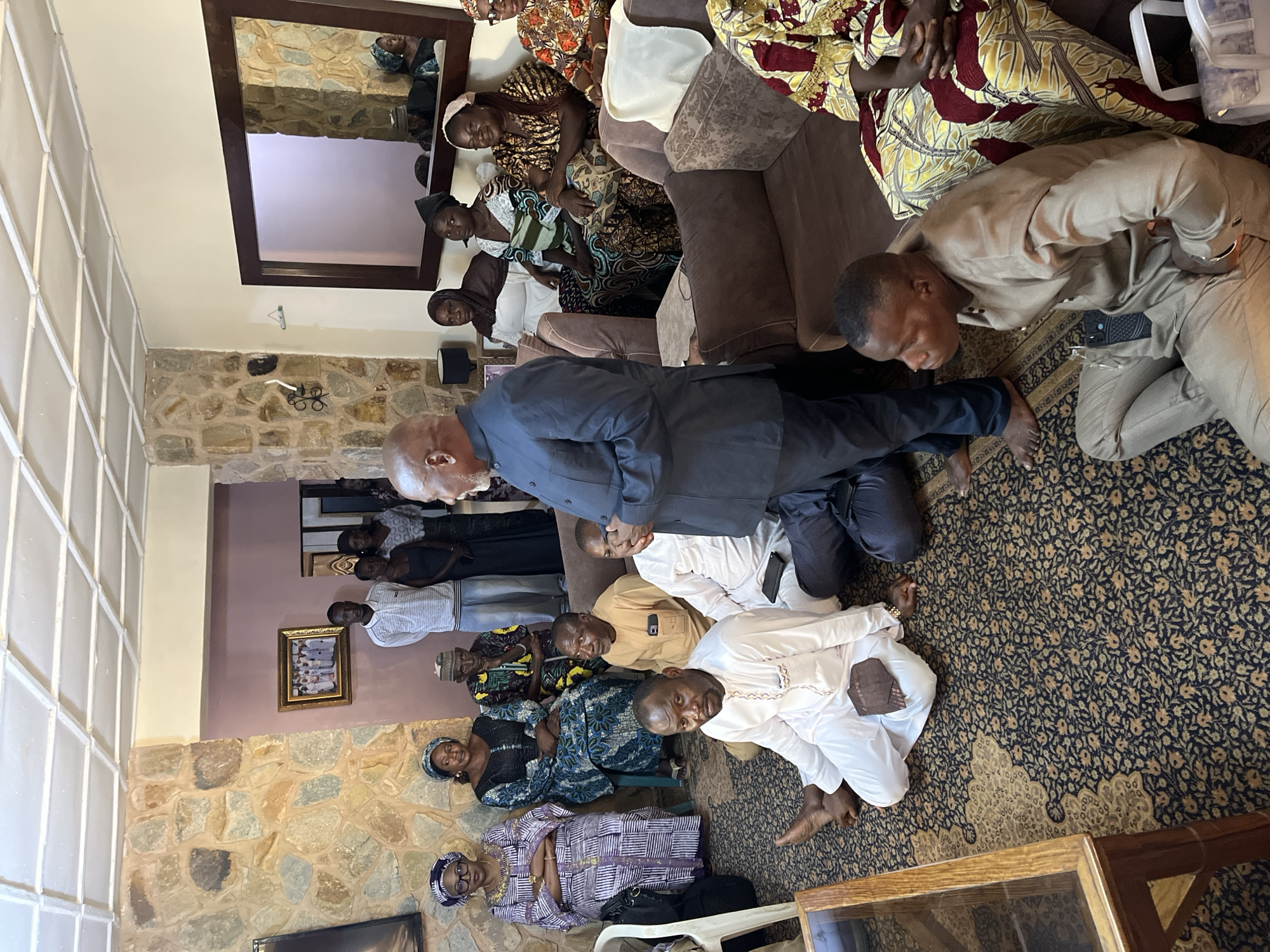 In Bukuru, Nigeria, a gathering takes place within a residential setting. The room is furnished with upholstered seating arranged around a patterned carpet. The walls are adorned with stone accents and a framed photograph. A large mirror hangs on one wall, reflecting the interior. Approximately ten individuals are present, some seated on the floor and others on sofas. They are dressed in traditional attire, suggesting a cultural event or social meeting. The atmosphere appears formal, with participants engaged in attentive postures. A man in a blue suit stands and addresses the group, while others listen.