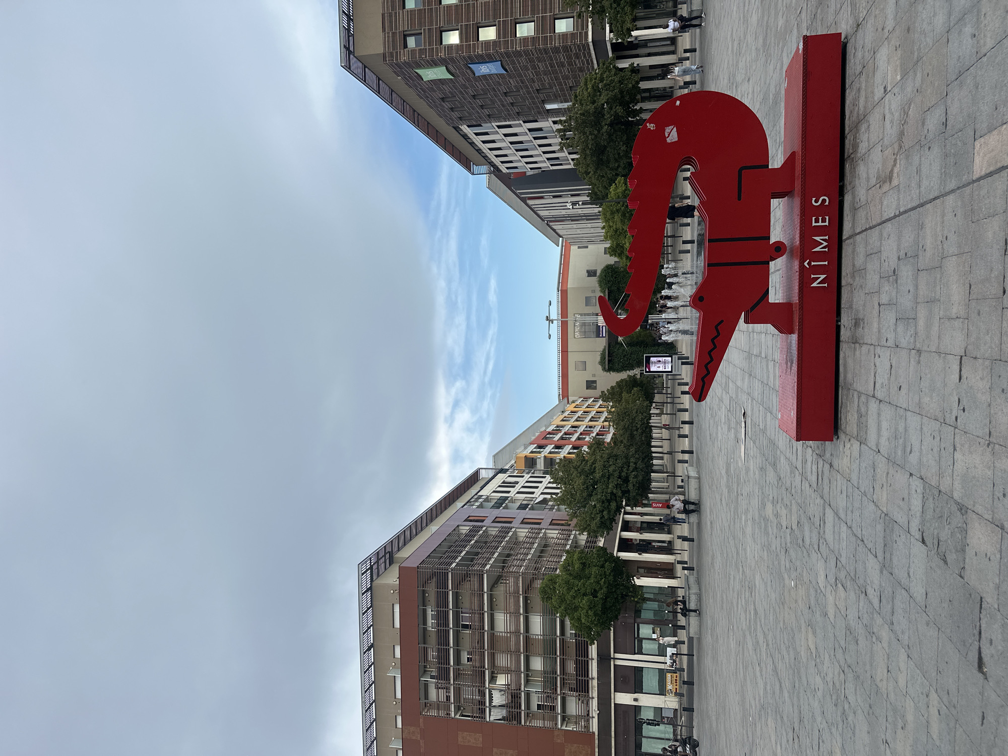 The image depicts a public square in Nîmes, France. In the foreground, a bright red, stylized alligator sculpture stands on a matching red base. The base is inscribed with the word "NÎMES" in white capital letters. Behind the sculpture, modern buildings with repeating window patterns and balconies line the square. Trees are planted along the walkway in front of the buildings. Several people are visible in the square, some sitting on benches, others walking. In the background, a fountain is active, with water jets visible. The sky is overcast with a mix of clouds.