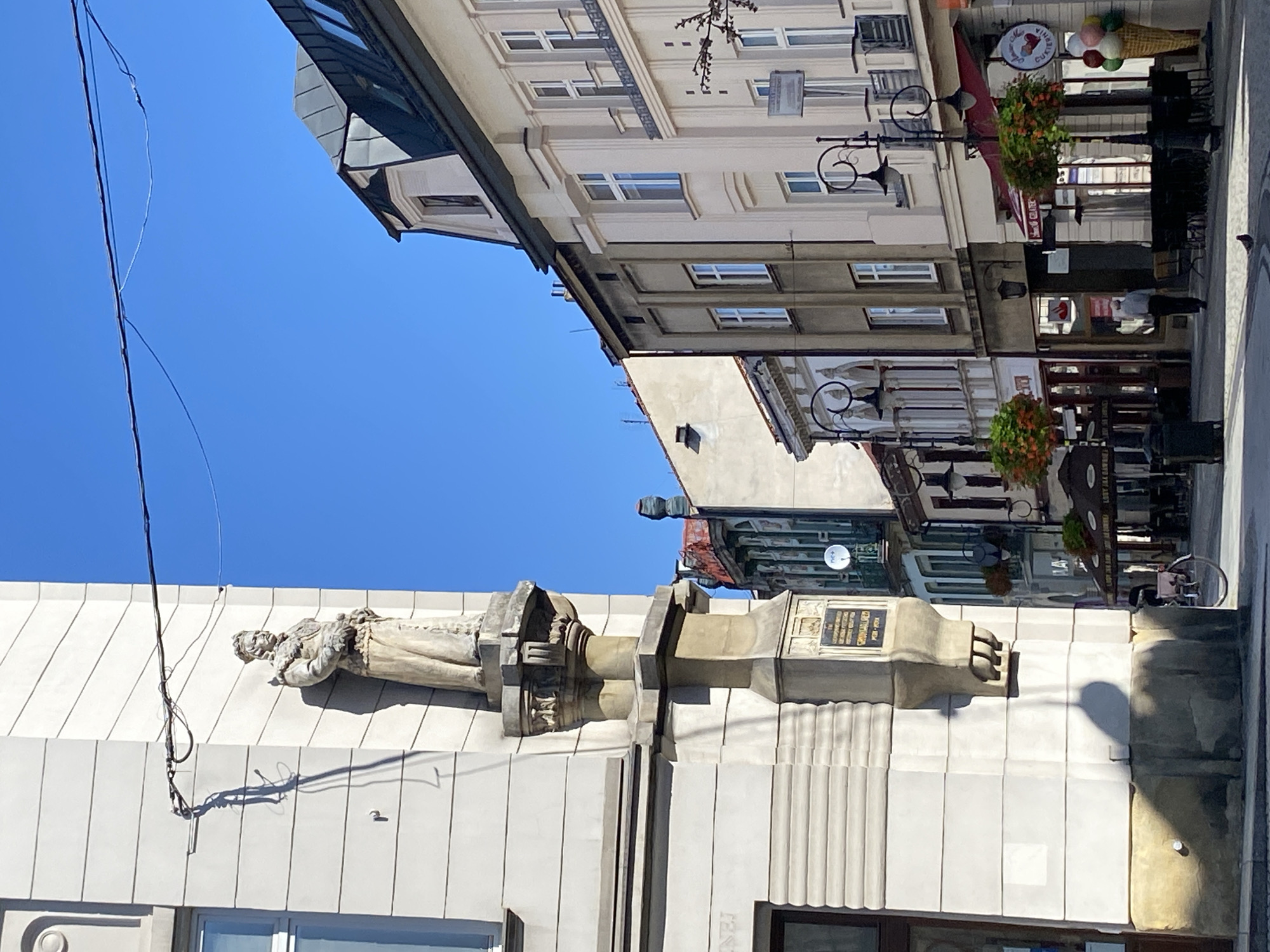 The image depicts a street scene in Nowy Sącz, Poland. On the left, a pale building features a prominent stone statue of a man in historical attire, wearing a crown and draped robes, mounted on an ornate pedestal. The statue casts a distinct shadow. Below the statue, a plaque is visible on the building's facade. To the right of the statue, the street continues with a row of buildings, characterized by their architectural styles and multiple windows. Some buildings have awnings and signs, suggesting commercial establishments such as cafes or shops. Street lamps with hanging flower baskets are positioned along the sidewalk, and outdoor seating areas with umbrellas are visible in front of some establishments. A bicycle is parked on the sidewalk, and a lone pigeon is on the cobblestone street. The sky is clear and blue, indicating fair weather. The overall impression is of a well-maintained urban street with historical elements.