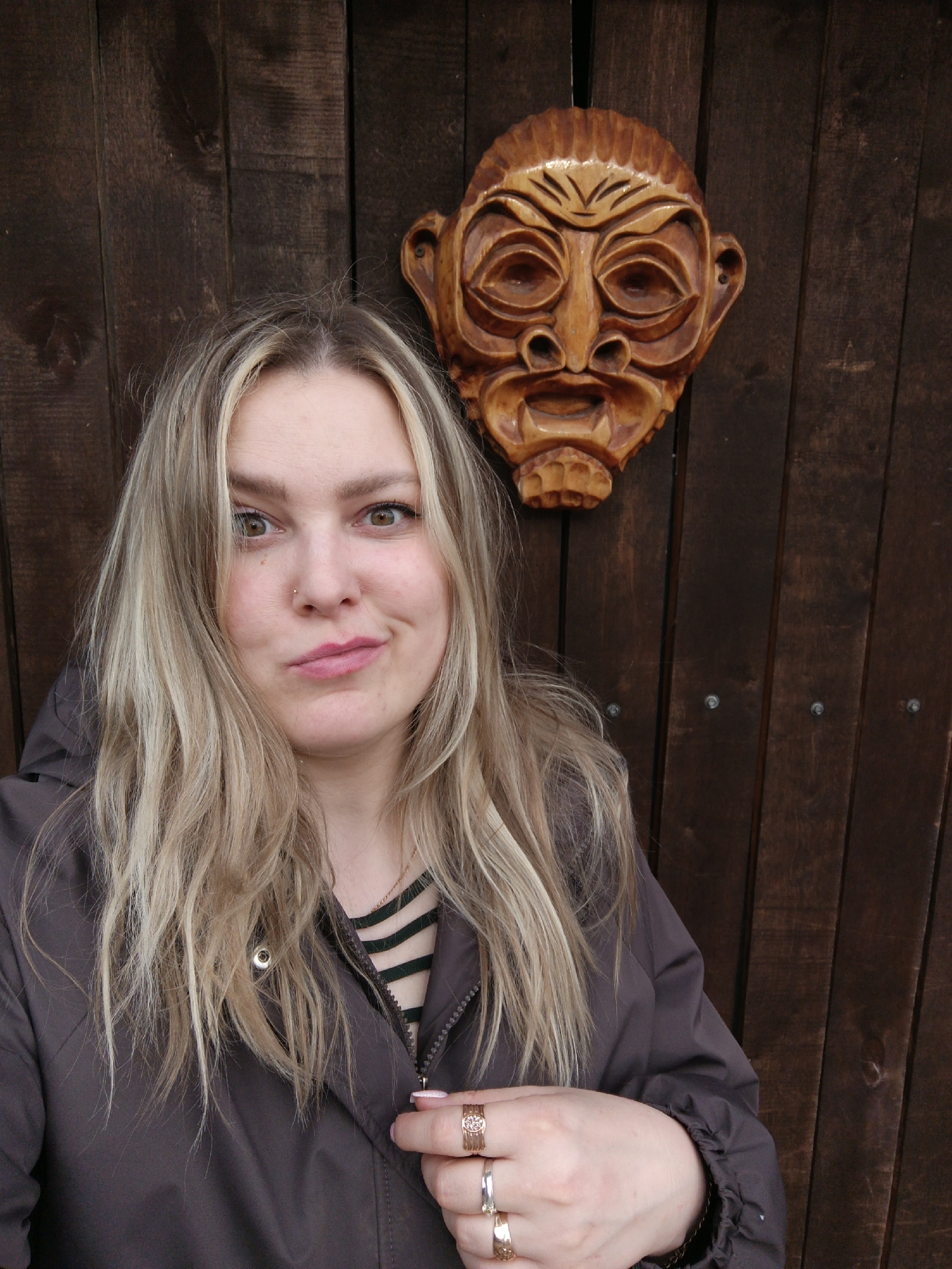 A woman poses for a portrait against a dark brown wooden plank wall in Chaikovsky, Russia. She looks directly at the camera with a playful, slightly questioning expression, her lips gently pursed. Her eyes are brown, and she has a small piercing on her left nostril. Her shoulder-length, wavy hair is blonde with visible darker roots, framing her face.
She is wearing a dark grey or brown hooded jacket, which is partially unzipped, revealing a glimpse of a dark green and black striped shirt underneath. Her right hand rests near the jacket's zipper, adorned with several rings: a wide, intricate golden ring, a silver ring, and another golden ring with a design. Her fingernails are painted a light pink.
Above and to her right, mounted on the wooden wall, is a carved wooden mask. The mask is light brown with darker, deeply carved details, depicting an expressive face with prominent brow ridges, wide, somewhat hollow eyes, a broad nose, and an open mouth. Its style appears folkloric or tribal. The wooden wall itself consists of vertical planks, with visible screws in some sections, suggesting a rustic outdoor setting. The lighting is soft and even, indicative of daytime, possibly an overcast sky. There is no visible text in the image.