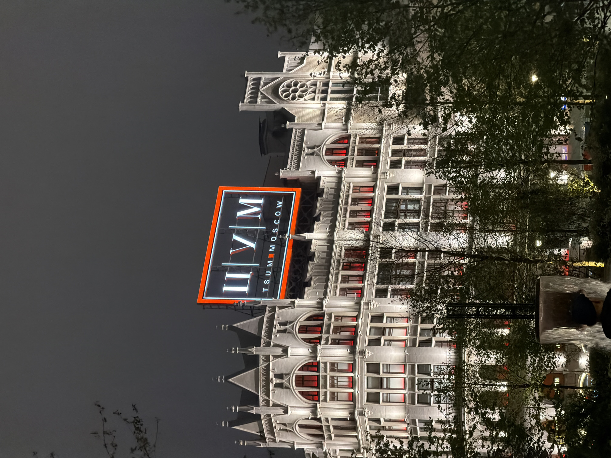At night, a tall, light-colored building with neo-gothic architectural elements stands prominently. A large, vertically oriented sign reading "TSUM MOSCOW" is affixed to the building. The building has numerous windows, with red lighting visible in some of them. Tree branches partially obscure the view of the building. The sky is dark and overcast. The scene is in Moscow, Russia, and features the TSUM department store.