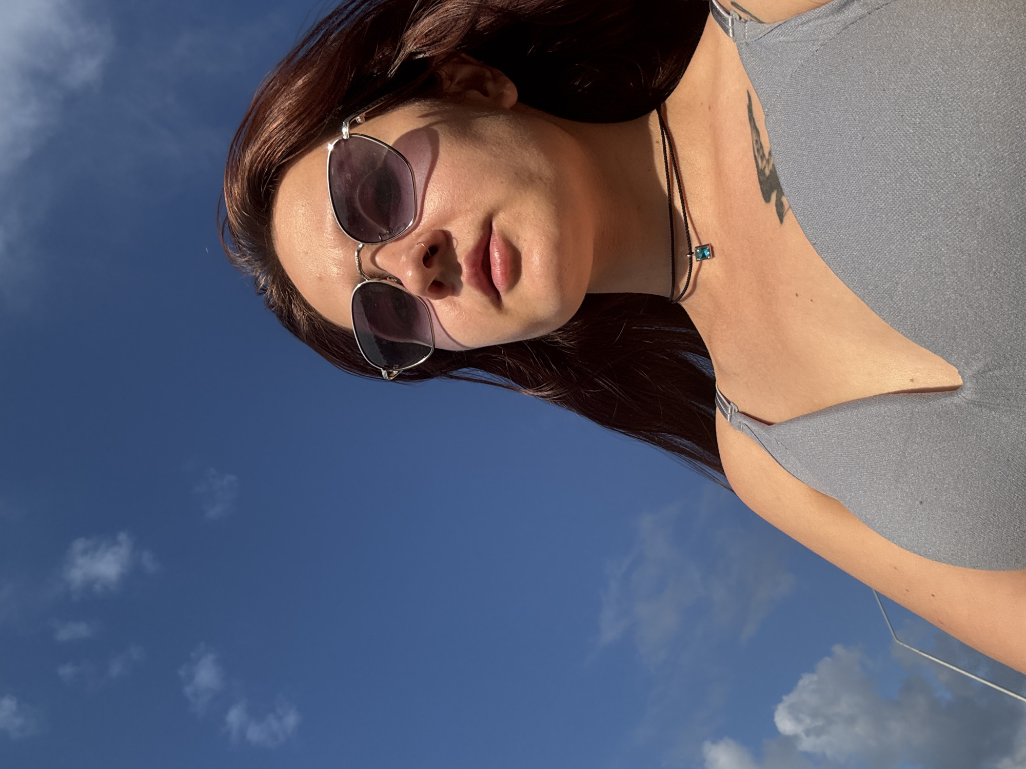 A young woman with long, dark auburn hair is photographed from a low angle against a bright blue sky with scattered white clouds. She is wearing sunglasses with silver frames, a thin black choker with a small, blue pendant, and a grey tank top. A tattoo is visible on her upper chest. The image appears to have been taken outdoors, possibly in Naberezhnye Chelny, Russia, given the bright, clear sky.