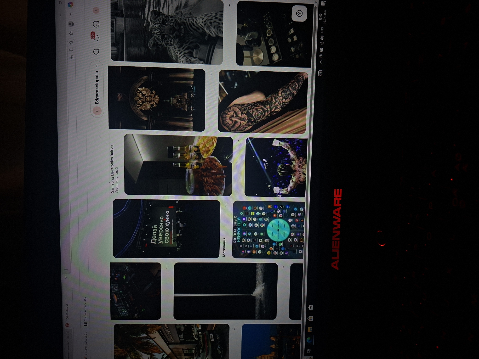 The image shows a close-up of a computer monitor displaying a Pinterest board. The Pinterest board features a grid of images, including a man in a boxing ring, a full sleeve tattoo of a dove and roses, a collection of watches, a leopard, a waterfall, and a group of bottles and snacks. The monitor is an Alienware brand, indicated by the red logo. The bottom of the screen shows the Windows taskbar with the time and date "13:09 19.07.2026". The background is dark and out of focus. There is no visible indication of the city of Užuežerė, Lithuania in the image.
