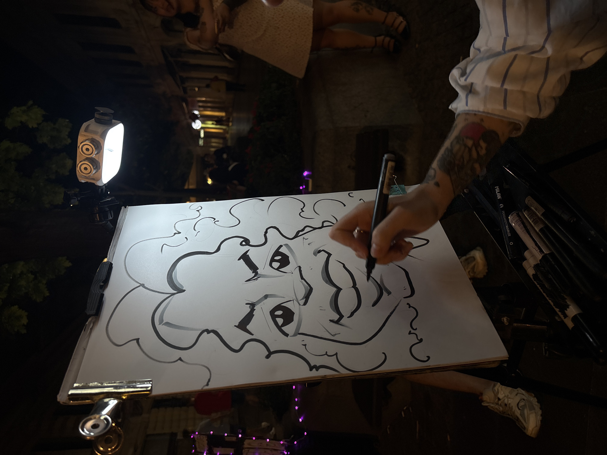 The image depicts a nighttime scene on a city street, possibly in Belgrade, Serbia, where a caricature artist is drawing. A large white board on an easel displays a black-and-white caricature of a face with large eyes, full lips, and curly hair. The artist's hand, adorned with tattoos on the forearm, holds a black marker and is in the process of adding details to the drawing. A spotlight, mounted on the easel, illuminates the drawing board and the artist's work.
In the background, the street is lined with buildings, some with lit windows, suggesting shops or restaurants. Several people are visible walking on the street, their forms blurred by the low light and focus on the foreground subject. Some purple string lights are strung along the left side of the image, adding a festive ambiance. The overall environment suggests a lively, pedestrian-friendly area during the evening. The artist is wearing a striped shirt, and a box of markers is positioned at the base of the easel. One person's legs, wearing a patterned dress and sandals, are visible on the right side of the frame, suggesting they might be a patron or passerby.