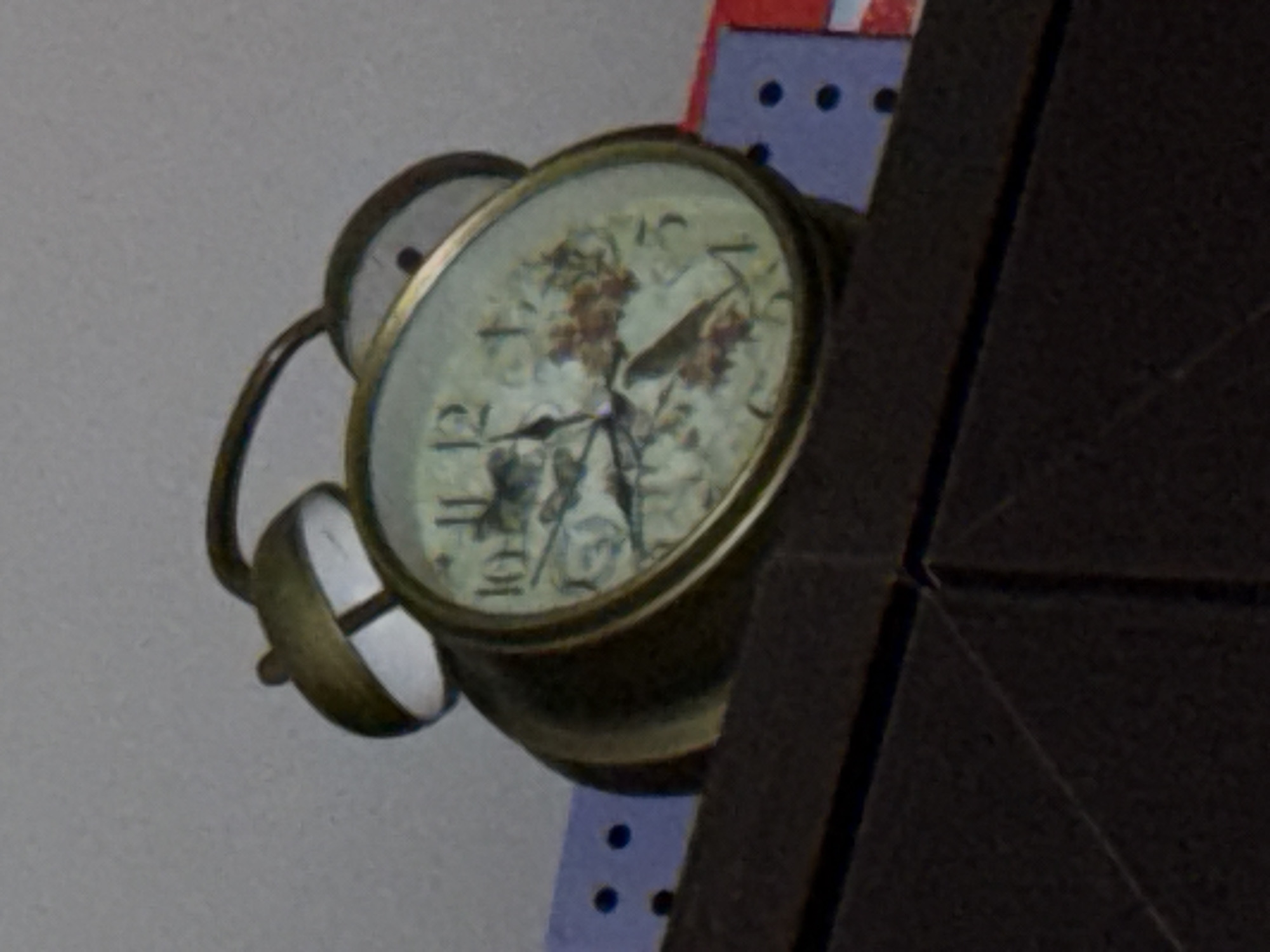 A close-up, low-angle shot shows a vintage alarm clock with a decorative face depicting a bird and flowers. The clock has a brass frame and two bells on top. It is positioned on the edge of a dark brown surface, possibly a shelf or table. In the background, a light gray wall is visible, with a section of blue and purple patterned material with black dots to the left of the clock, and a red and blue striped material to the right. The clock's hands indicate a time close to 10:55.