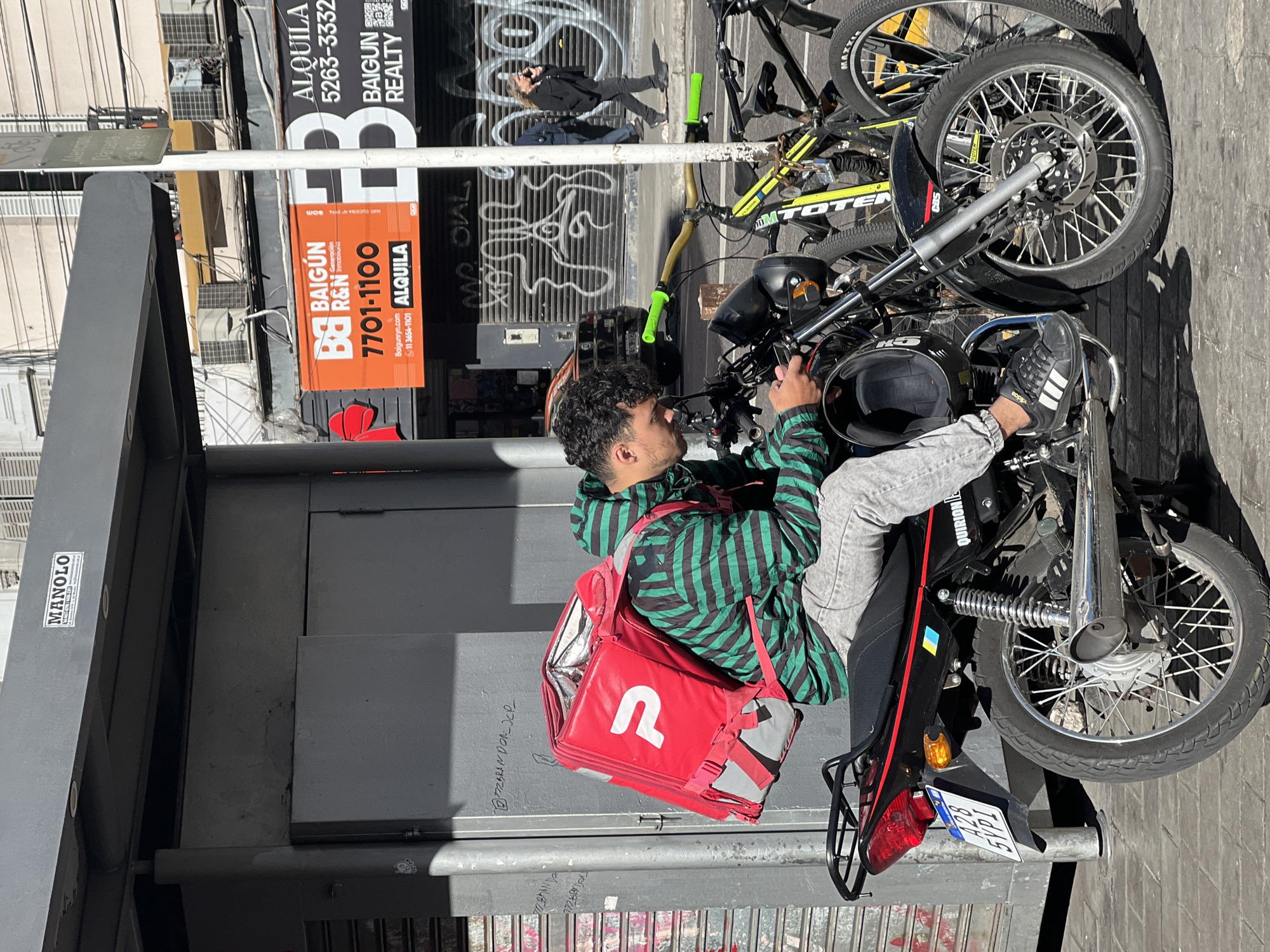 The image shows a delivery man sitting on a black motorcycle in Buenos Aires, Argentina. He wears a green and black striped jacket, gray pants, and black Adidas sneakers. A red delivery backpack with a white "R" logo sits behind him. The motorcycle has a license plate with the number A28 and the inscription QUIRION. A black helmet rests on the motorcycle's handlebars. Another black and yellow bicycle is parked nearby.
Behind the motorcycle, graffiti is visible on the wall of a building. Above the graffiti, signs for "BAIGUN REALTY" and "ALQUILA" are displayed with the phone numbers 7701-1100 and 5263-3332. A bus stop shelter with a "MANOLO" sticker is to the left of the delivery man.