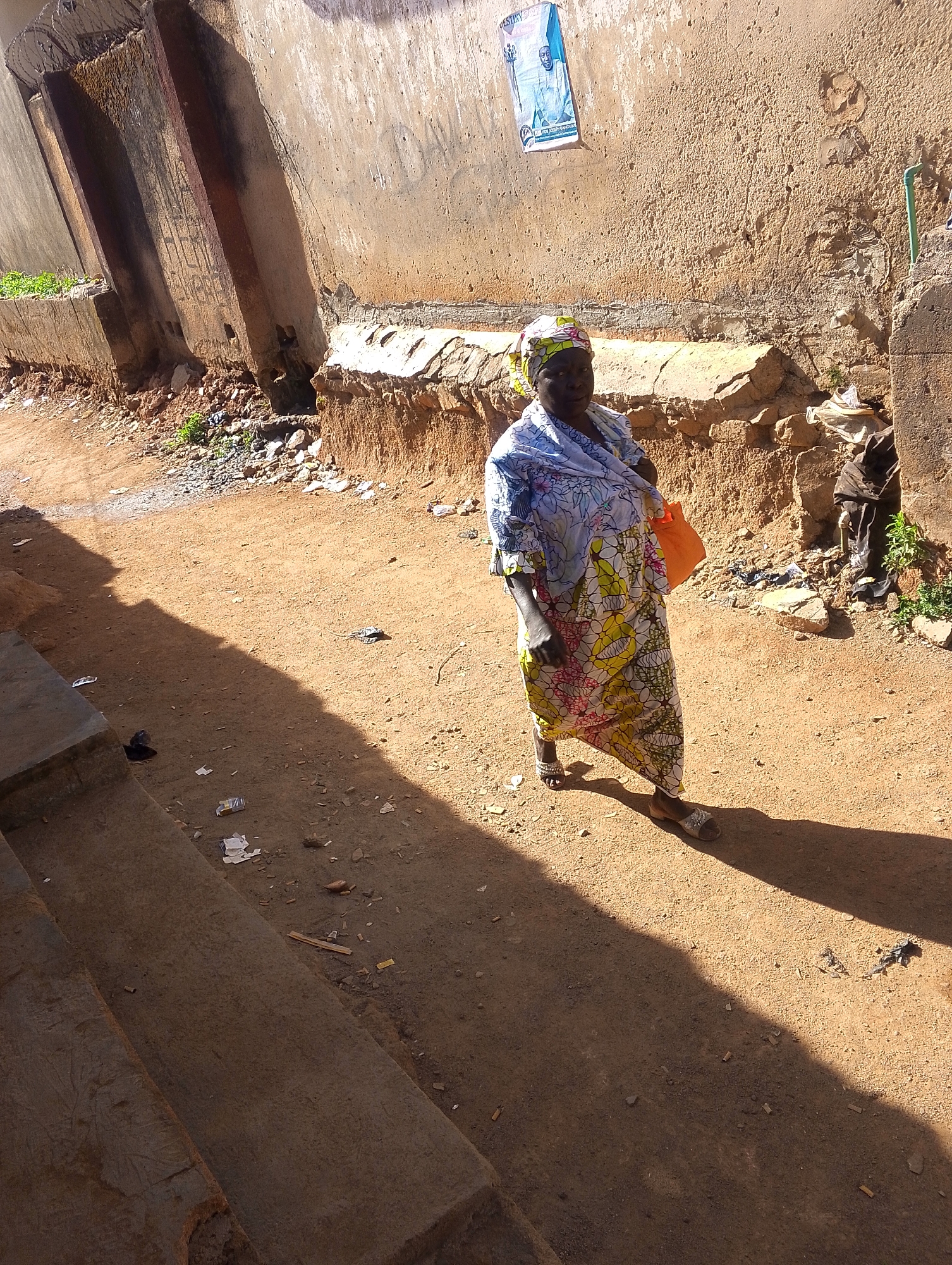 A woman in a floral dress and headscarf walks on a dirt road in Jos, Nigeria. She carries an orange bag. A poster hangs on a weathered wall. The ground is littered with debris. Buildings border the street.