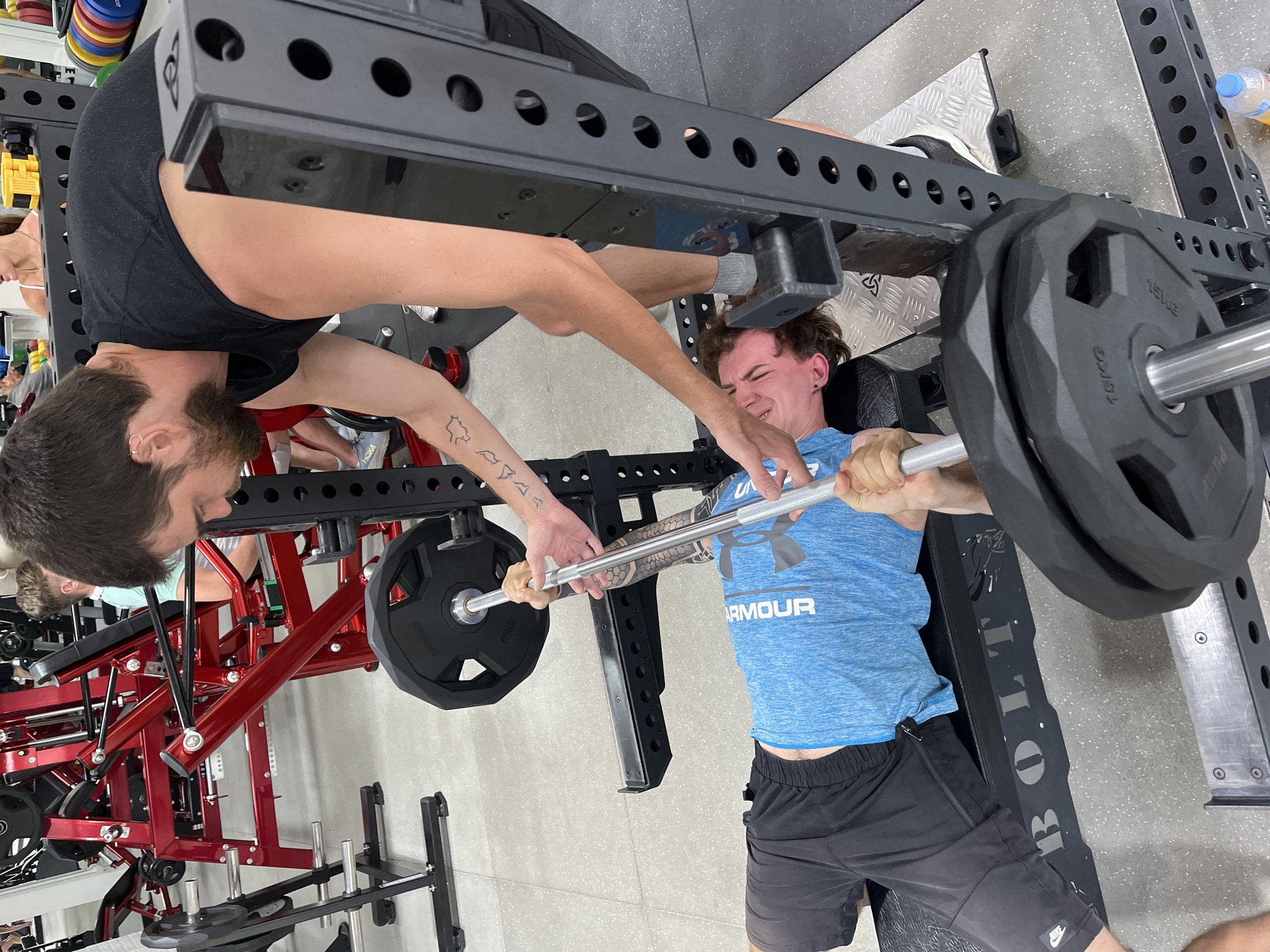 This image captures a scene inside a gym, likely located within Chiang Mai City Municipality, Thailand. The primary action involves two individuals engaged in a weightlifting exercise, specifically a bench press.
In the foreground, a young man lies supine on a black weight bench, wearing a blue Under Armour t-shirt and black shorts. He is actively performing a bench press, gripping a barbell loaded with black weight plates. His face is contorted with exertion, indicating significant effort.
Behind him, another individual, a man with dark hair and a beard, stands and assists with the bench press, his hands positioned near the barbell to provide support and spot the lifter. He is wearing a black t-shirt.
The gym environment is characterized by various weightlifting equipment. To the left, red weight machines are visible, and to the right, a black squat rack is prominent. The floor appears to be a light gray speckled surface. The overall lighting suggests an indoor, well-lit gym. The presence of other gym-goers and equipment in the background suggests a typical gym setting.
