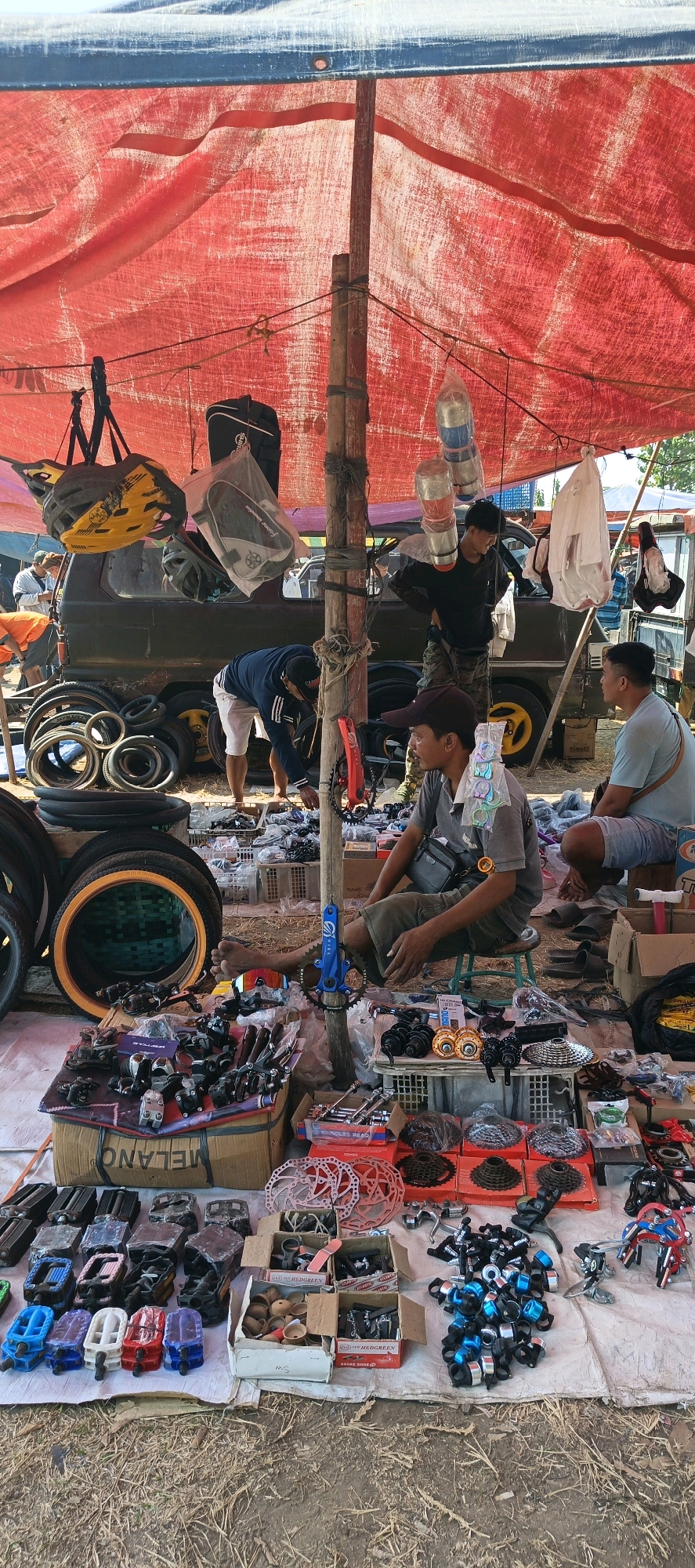 In Jatinom, Indonesia, an outdoor market stall displays a variety of bicycle parts and accessories under a large red tarp. The ground is covered with dry grass and dirt. Three men are visible. In the foreground, a man wearing a dark cap and a gray t-shirt sits on a stool, looking to his right. To his right, another man in a light blue t-shirt and shorts is seated, also looking to his right. Further back, a man in a dark blue shirt and white shorts is bent over, examining items on the ground. The stall is laden with bicycle tires, brake discs, gears, pedals, and various metal components, some of which are arranged in boxes and on a cardboard surface. A dark-colored van is parked behind the stall, and other market activity is suggested in the background. The scene depicts a bustling marketplace for bicycle parts.