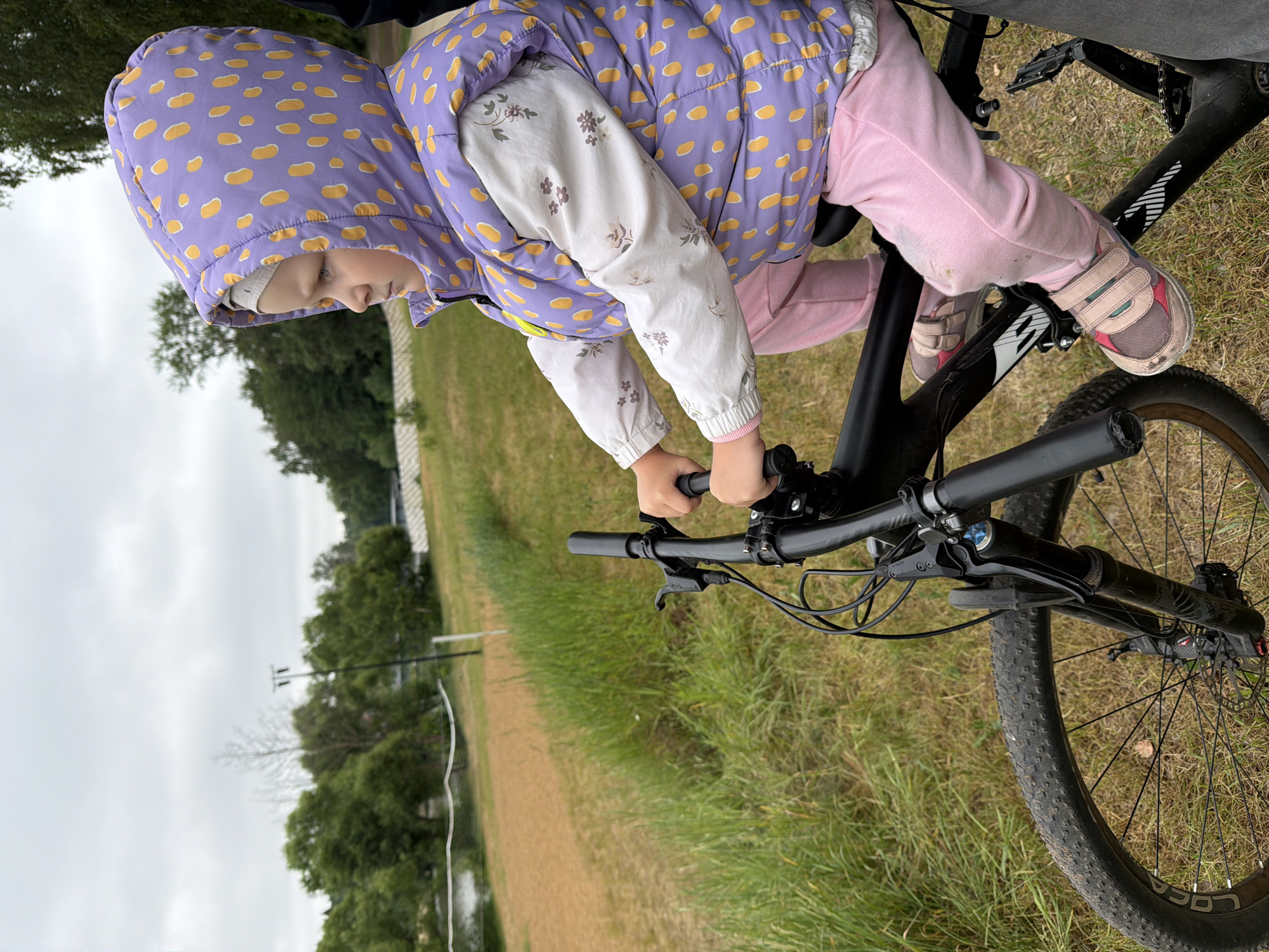 A young child wearing a purple hooded vest with yellow polka dots and pink pants is sitting on a black bicycle. The child's hands are on the handlebars, and they are looking to the right. The bicycle is on a grassy area next to a dirt path and a body of water. In the background, there are trees and a cloudy sky. The scene is likely located in Malakhovka, Russia, given the context. The child is facing away from the camera and appears to be engaged with the environment around them, possibly on an outing or adventure. The overall atmosphere is one of outdoor activity in a natural setting.