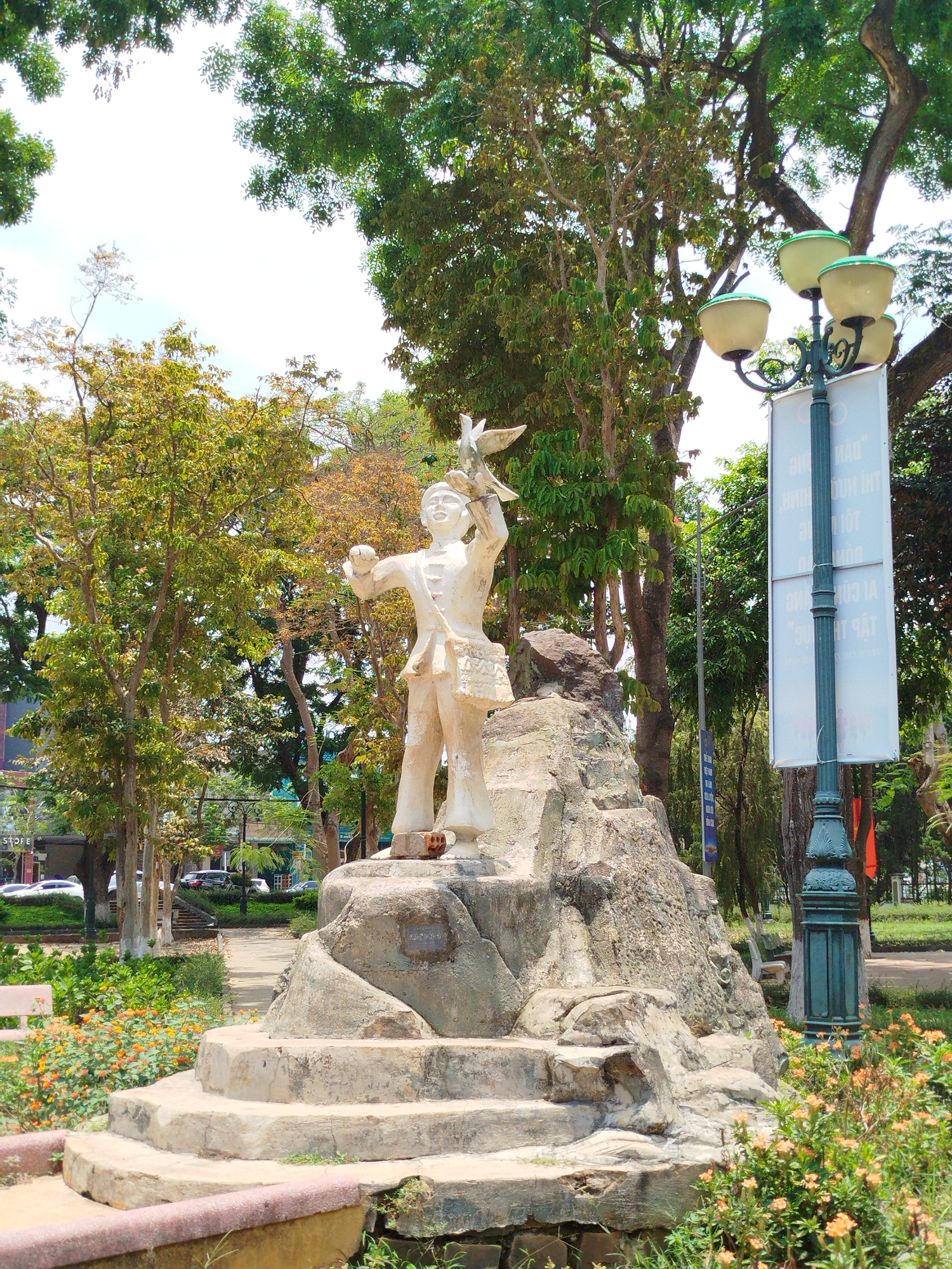 A statue of a person is located on top of a multi-tiered stone plinth surrounded by flowers and trees. The statue depicts a figure in traditional clothing with a bird in their hand, seemingly releasing it into the air. The statue is made of a light-colored material and appears to be weathered. The surrounding vegetation consists of trees with a variety of green and brown foliage. A lamp post with multiple globe lights stands near the statue. A banner is attached to the lamp post. The scene is likely located in the town of Liên Nghĩa, Vietnam.