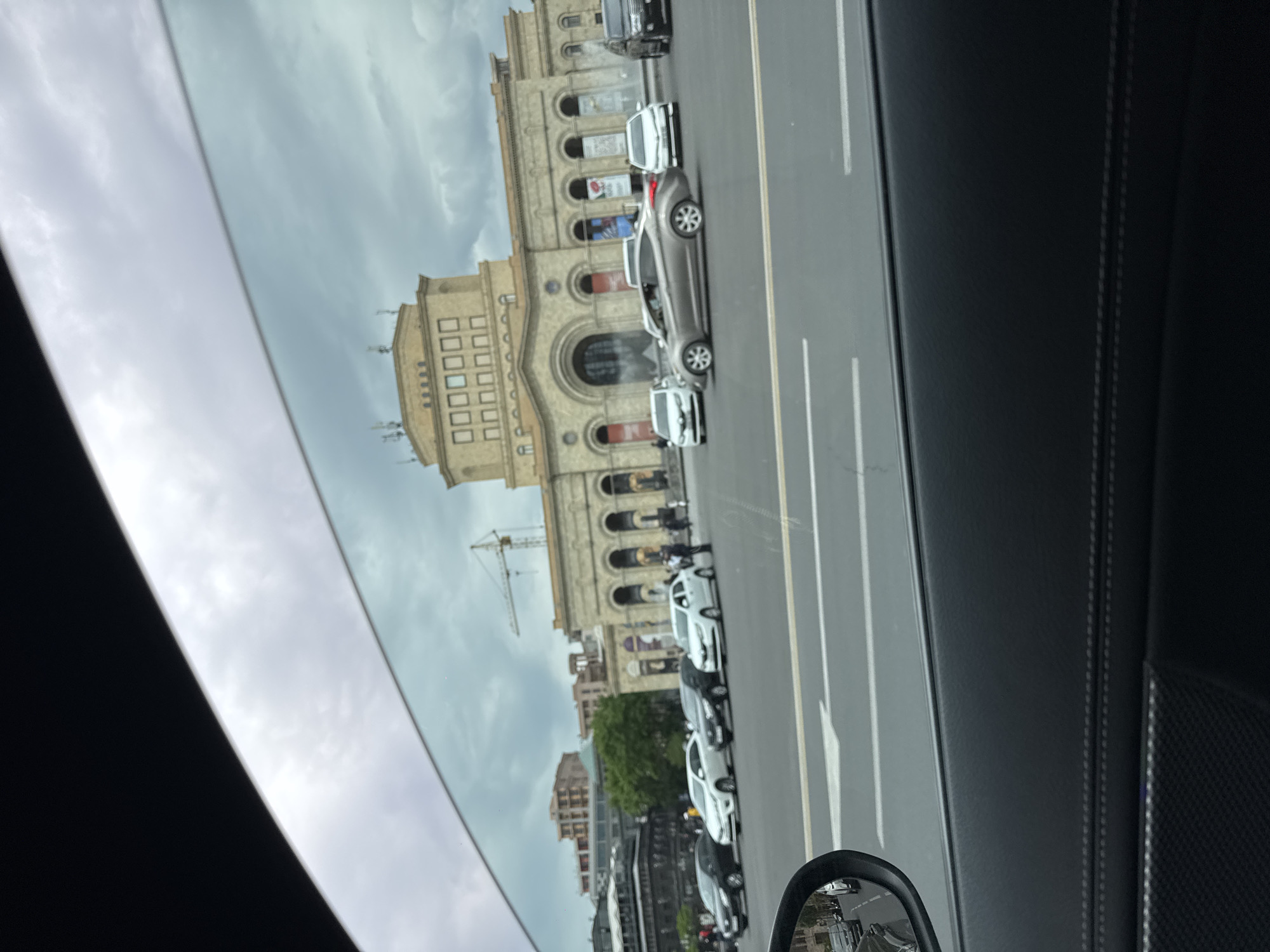 The image, taken from inside a vehicle, shows Republic Square in Yerevan, Armenia. Visible is the Government House building. Cars are parked along the side of the road. A construction crane is visible in the background. The sky is overcast. The vehicle's window frame and side mirror are also visible in the foreground.