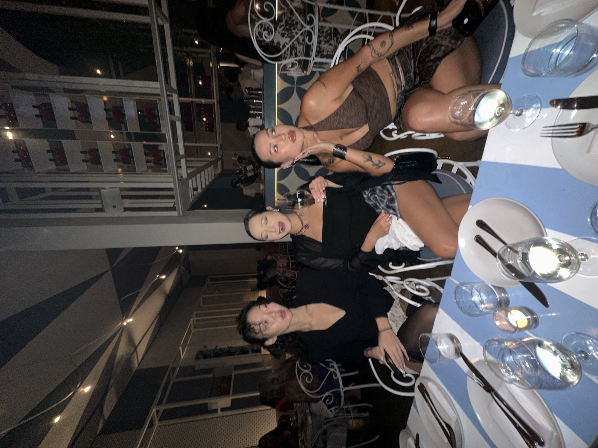 The image depicts a nighttime scene inside a restaurant, potentially located in North Kuta, Indonesia. Three young women are seated at a table set for a meal. The woman on the left wears a black jacket and a patterned skirt, while the woman in the center is in a black top and a leopard-print skirt. The woman on the right is wearing a brown halter top and a matching skirt. They appear to be conversing and enjoying drinks. The restaurant's interior features a decorative ceiling with diagonal stripes and hanging lights, along with shelves displaying bottles in the background. The table is adorned with white plates, cutlery, glasses, and a small candle. The overall ambiance suggests a social gathering in a well-lit, upscale dining establishment.