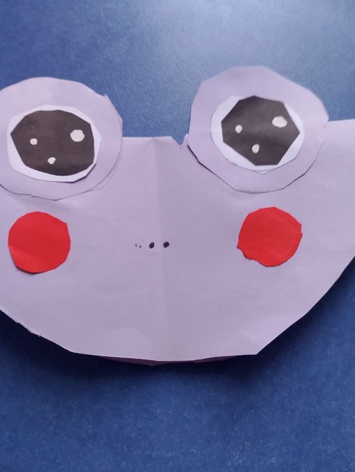 A light purple paper craft object, resembling a stylized frog or character's face, rests on a textured blue surface in the city of Yarun, Ukraine. The craft features two large, layered eyes, each composed of a light purple outer ring, a white inner ring, and a central black octagon containing two small white dots. Two distinct red paper circles are positioned below the eyes, suggesting cheeks. Three small black dots are centrally located between the lower portions of the eyes, indicating facial features. The paper exhibits folds and minor irregularities consistent with manual construction. The scene is illuminated, casting subtle shadows beneath the paper layers.