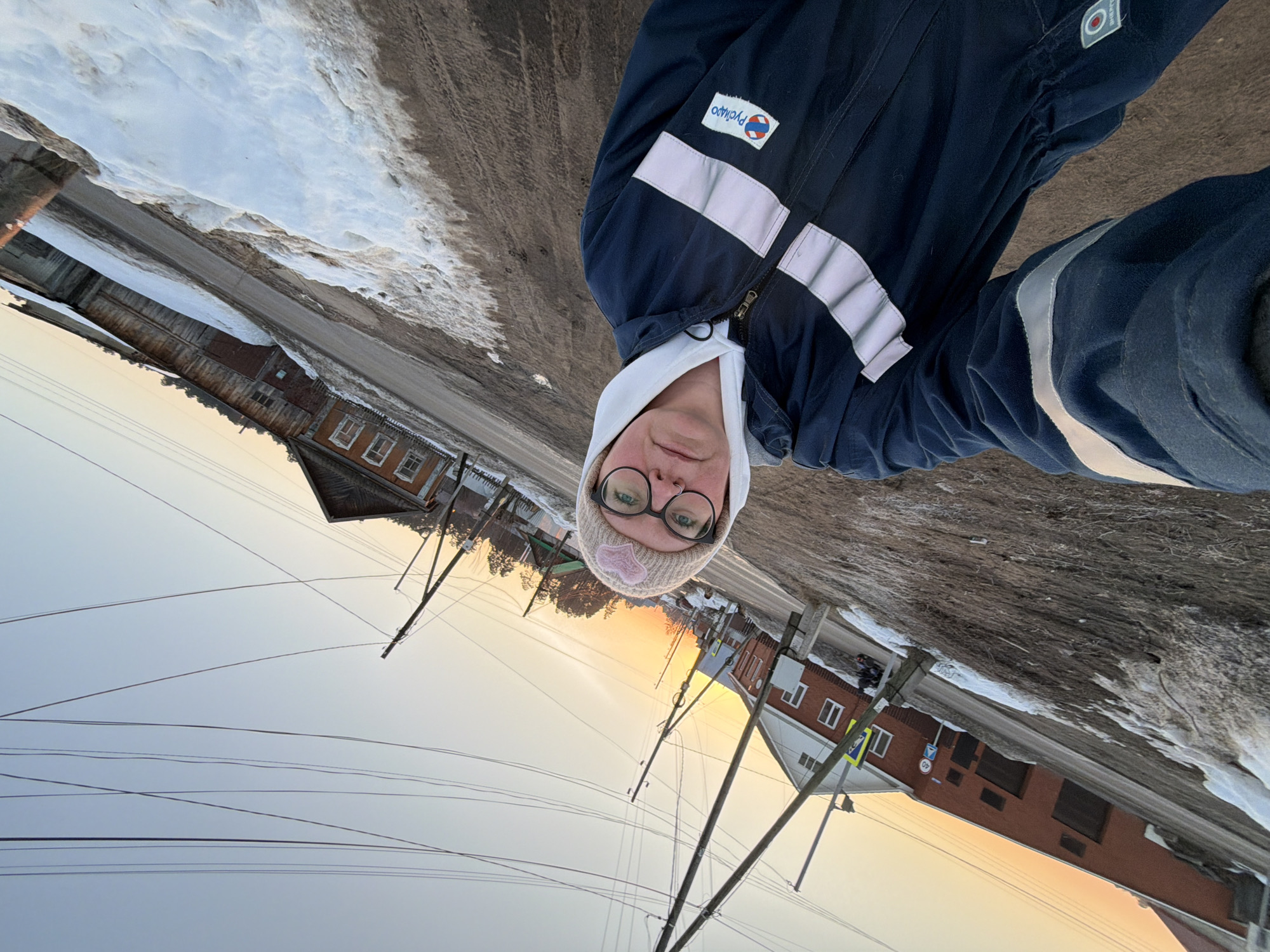 The image presents an outdoor selfie taken on a street in Chaikovsky, Russia, at either sunrise or sunset, indicated by the warm, low-angle light. A single individual is visible in the foreground, positioned slightly right of center, taking the photograph. This person wears a dark blue work jacket featuring two light grey reflective stripes across the chest and one on the right sleeve. A white hooded sweatshirt is visible underneath the jacket. An embroidered "РусГидро" (RusHydro) logo is present on the left chest, and a circular logo is on the lower right sleeve. The individual wears dark-rimmed glasses and a light-colored knit beanie with a pink heart emblem on the front.
The background depicts a residential street environment. The road surface is unpaved, composed of dirt and mud, with visible tire tracks. Patches of melting snow and ice line the sides of the road. Numerous wooden utility poles with multiple overhead power lines extend across the scene against a sky that transitions from a pale orange-yellow at the horizon to a clear light blue overhead. On the left side of the road, a two-story red brick building with white window frames and a white gable roof is visible, near which a yellow triangular pedestrian crossing sign is mounted. Further into the distance, several older wooden houses with dark windows line both sides of the street. A tall, dark wooden fence runs parallel to the road on the right. Sparse, bare trees are visible on the horizon, suggesting a late winter or early spring season.