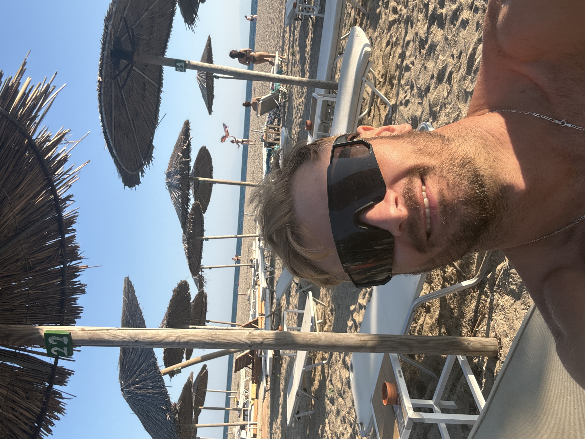 A man takes a selfie on a beach in Angelochori, Greece. He is wearing sunglasses and a silver chain necklace. Behind him, straw umbrellas shade rows of lounge chairs on the sandy beach. In the background, people are visible near the water, and a kite is flying in the sky. The scene is bathed in sunlight under a clear blue sky.