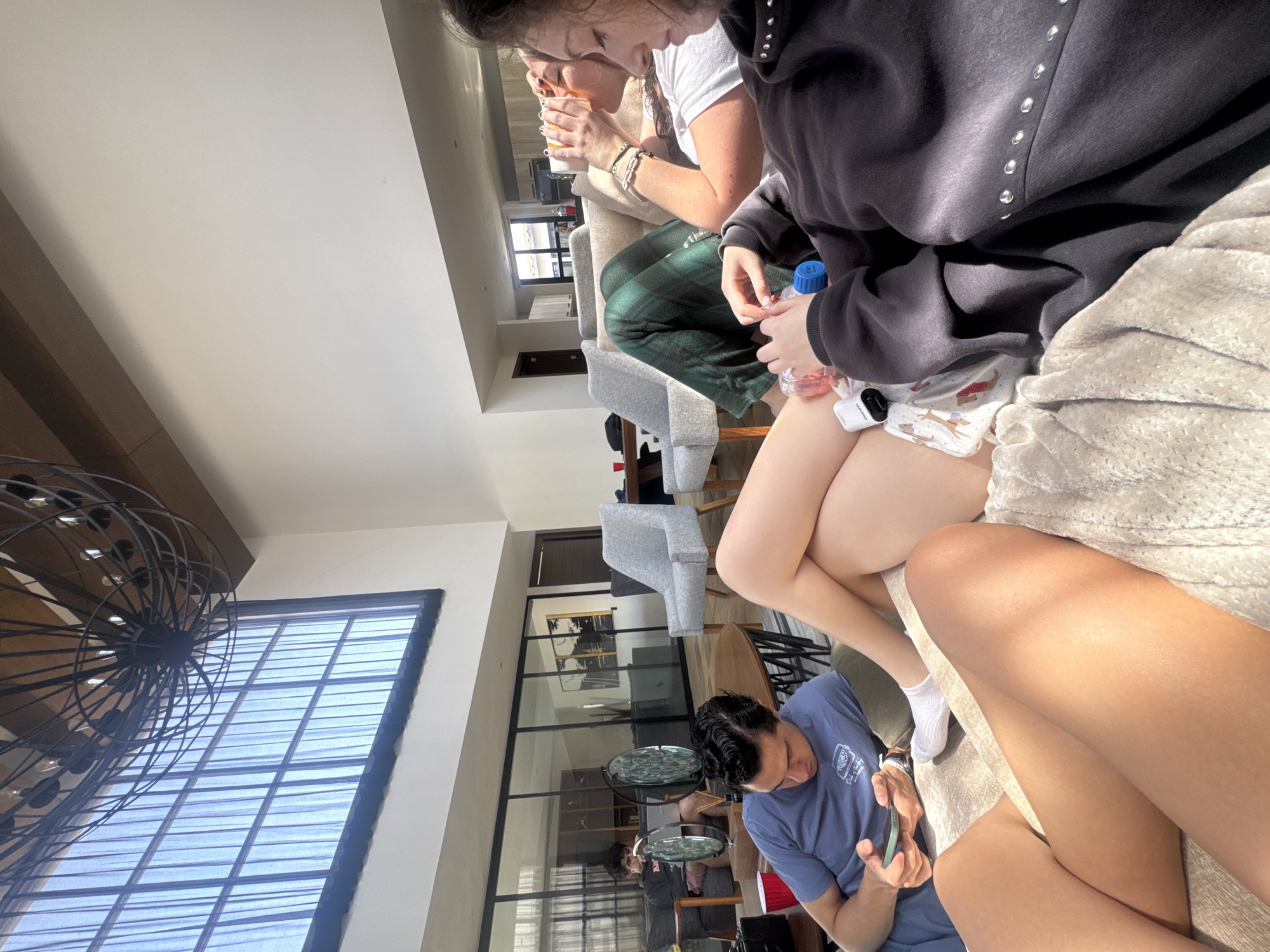 Here is a description of the image:
The photograph depicts a group of people in what appears to be an indoor setting, possibly a lounge or a common area. At the top of the image, one person is drinking from a cup, while another next to them is wearing a black hoodie with silver embellishments. The second person also has a water bottle. Near the bottom of the frame, a person wearing a blue shirt is using a smartphone. The background includes large windows, gray armchairs, and interior design elements. The location in the city of Unknown, Unknown is unconfirmed.