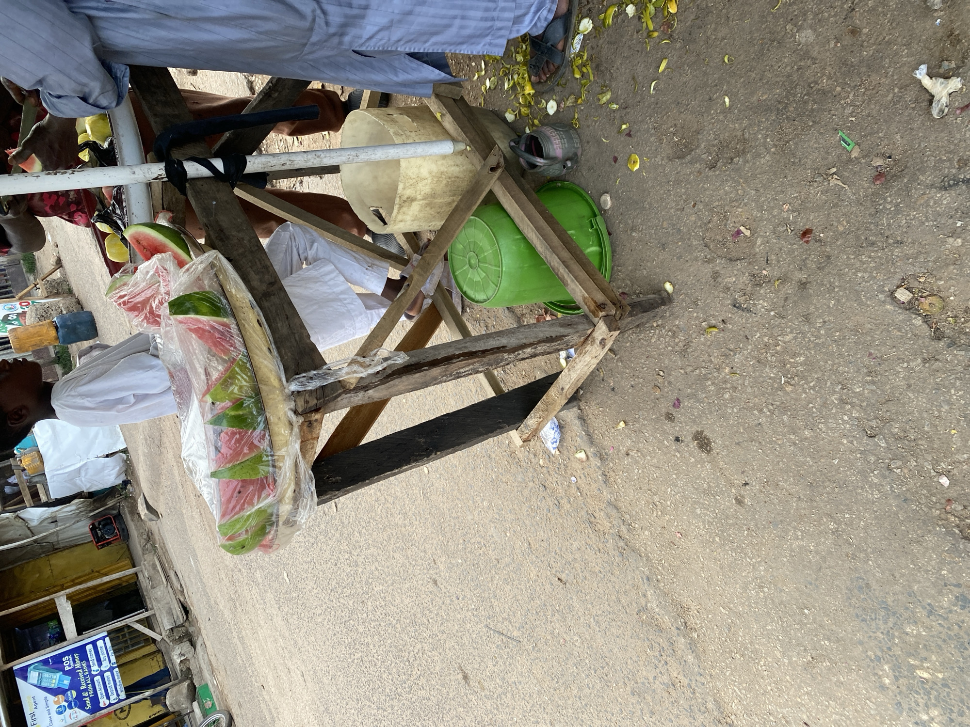 A street vendor in Jos, Nigeria, is preparing and selling sliced watermelon from a wooden stand on the side of a paved road. The vendor, a young person wearing a white tunic, is on the left side of the frame, with sliced watermelon pieces wrapped in clear plastic arranged on the stand. To the right, an adult, possibly another vendor or customer, is wearing a light blue robe and is in the process of cutting or handling a piece of fruit, likely watermelon, with a knife. Other individuals are visible in the background, some appearing to be involved in the transaction or simply present in the market area. Scattered on the ground around the stand are remnants of fruit peels, leaves, and other debris, indicating a busy street-side market environment. The background shows stalls and buildings, typical of an urban marketplace.