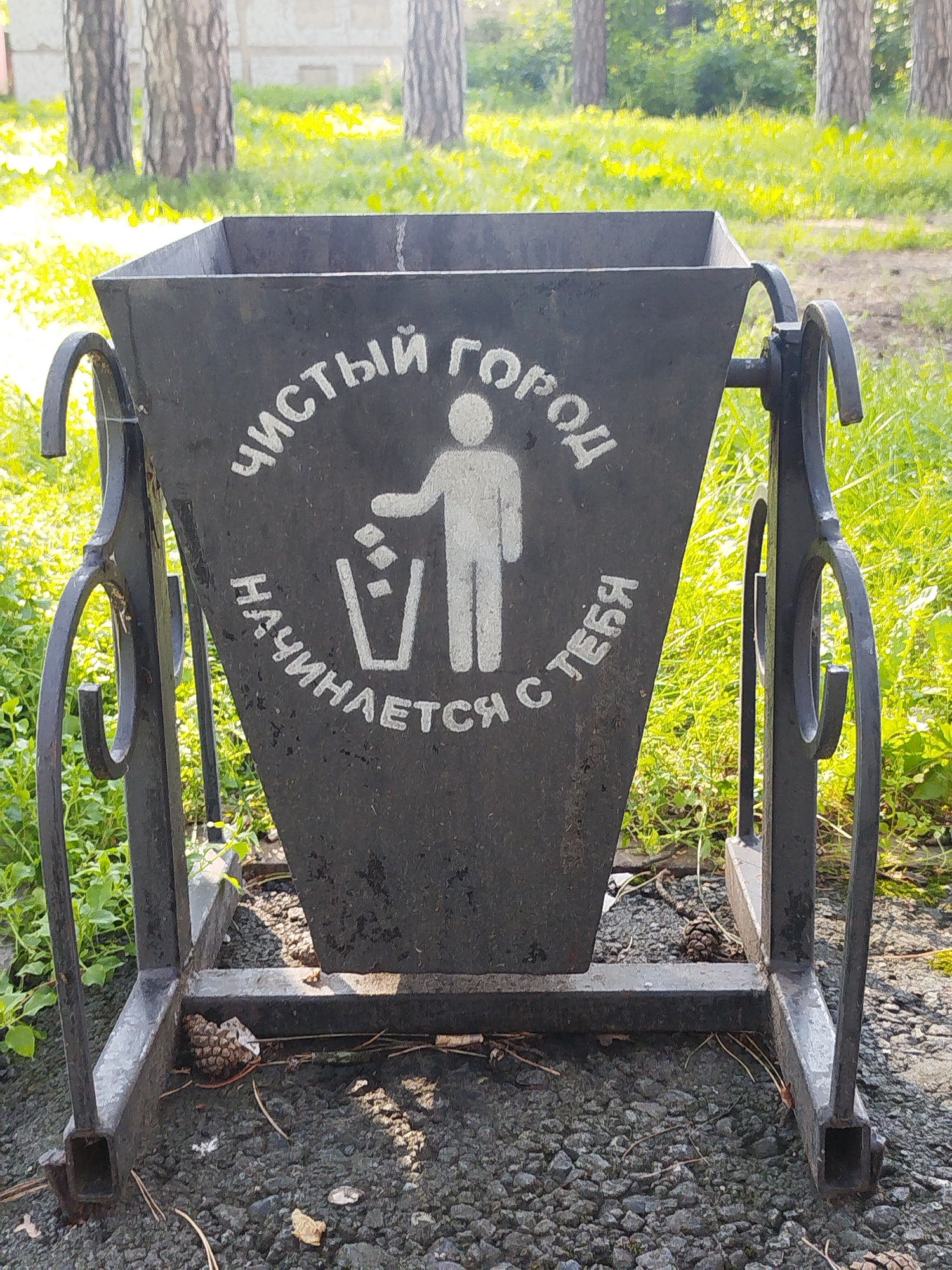 An outdoor scene features a dark metal trash receptacle with a conical-shaped body and an ornate wrought-iron stand. White text and a graphic of a person discarding an item into a trash can are visible on the front of the receptacle. The text reads "ЧИСТЫЙ ГОРОД НАЧИНАЕТСЯ С ТЕБЯ," which translates to "A clean city starts with you." The receptacle is situated on a gravel surface, with green grass and trees in the background. The setting appears to be a park or a public area in Zavodoukovsk, Russia, evidenced by the specific Cyrillic text on the trash can, commonly found in Russian public spaces, and the general landscape which is consistent with the region. The lighting suggests it is daytime, with sunlight illuminating the scene. The image is captured from a frontal perspective, at a medium distance, focusing on the trash receptacle and its immediate surroundings.
