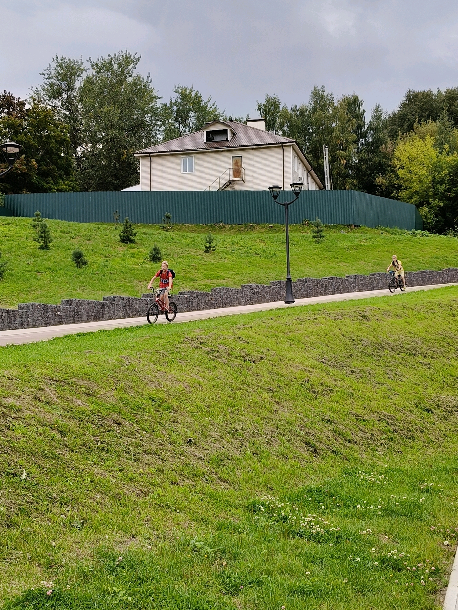 A young person with blonde hair, wearing a red t-shirt and shorts, is riding a bicycle on a paved path in a grassy park. Behind them, a second person, also with blonde hair, wearing a yellow top and shorts, is riding a bicycle on the same path. The path is lined with a stone wall on one side and a green fence on the other. In the background, there is a light-colored house with a dark roof, surrounded by trees. The sky is overcast. The scene appears to be in a park or recreational area in Naro-Fominsk, Russia, likely during daylight hours.