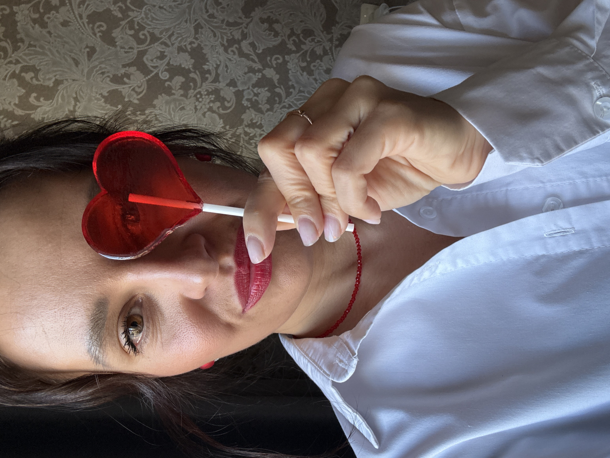 A woman with brunette hair is pictured from the chest up, rotated 90 degrees counterclockwise. She is wearing a white button-down shirt and a thin red beaded necklace. She is holding a red heart-shaped lollipop with a white stick in front of her left eye. She has a gold ring on her left hand. Her lips are painted a deep red. The background is a beige floral wallpaper. The location is Chaikovsky, Russia.