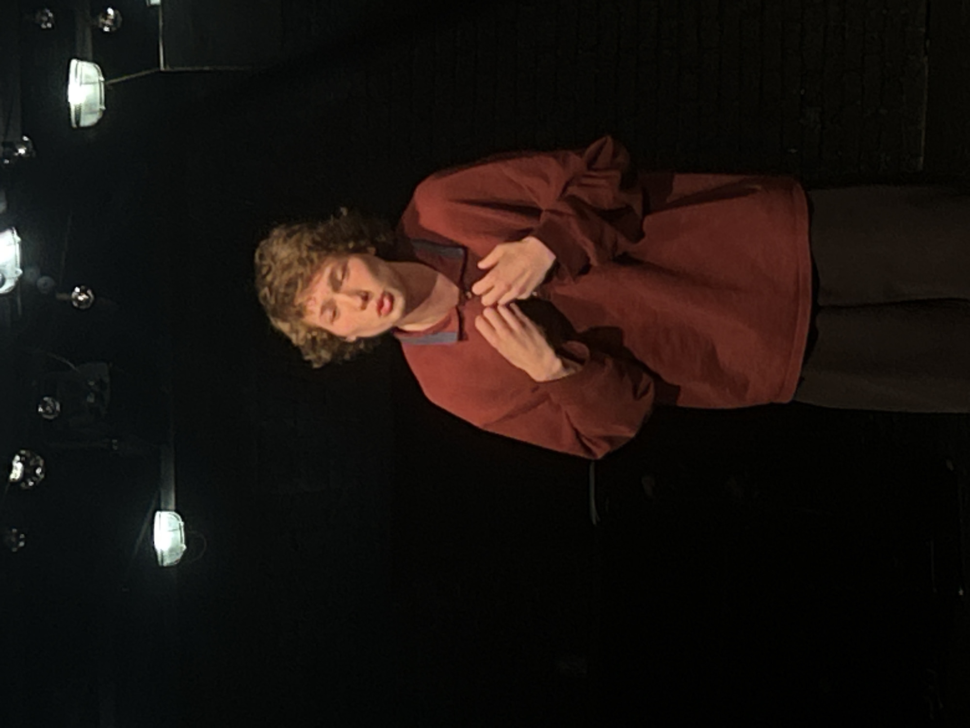A single person, appearing to be a young male with curly brown hair, is depicted in what seems to be a performance setting against a dark, featureless background. The individual is wearing a long-sleeved, collared shirt in a deep red or maroon color, with dark pants. Both hands are placed on their chest, near the collar of the shirt. Their mouth is slightly open, revealing a reddish hue on the lips. The expression on their face is somewhat dramatic or intense, possibly conveying emotion.
The background is predominantly dark, suggesting a stage or a darkened room with selective lighting. Several light fixtures, possibly stage lights or spotlights, are visible on the left side of the frame, appearing as bright, reflective, or illuminated objects against the blackness. The lighting is focused on the subject, highlighting them against the dark environment. No specific location cues beyond an indoor performance space are evident. There is no visible text. The overall scene conveys a sense of a staged performance or a posed, emotional moment.