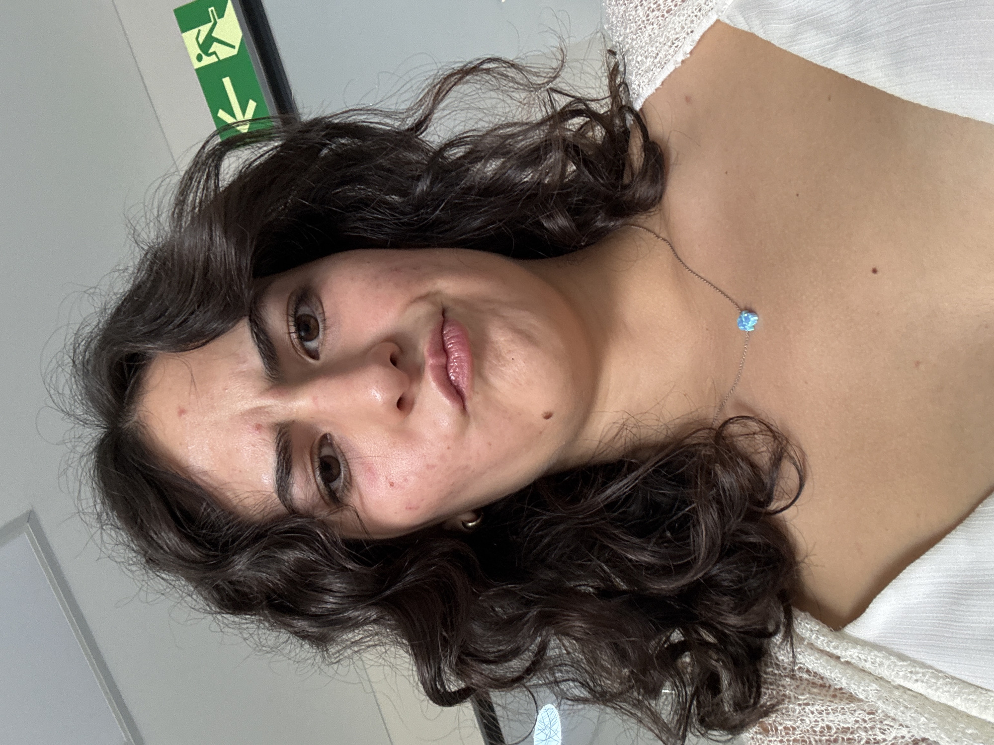 A close-up portrait features a young woman with dark, wavy or curly hair, looking directly at the camera with a subtle, slight smile. Her head is tilted to the left within the rotated image frame. She has light brown eyes, fair skin with some minor blemishes on her forehead and cheeks, and light pink lips. A small mole is visible to the right of her mouth.
She is wearing a light-colored, possibly white or cream, top with a textured, slightly sheer fabric visible on her right shoulder and décolletage. A delicate silver chain necklace with a small, light blue, heart-shaped pendant, possibly made of opal, rests on her chest.
The background consists of a light grey wall. In the upper left corner of the image, a green emergency exit sign is visible. The sign displays the universal symbol of a running person and a downward arrow.
The setting appears to be indoors, likely a public or commercial building in San Andrés Cholula, Mexico, as suggested by the emergency exit sign. The image captures a moment, possibly a selfie or a casual portrait.