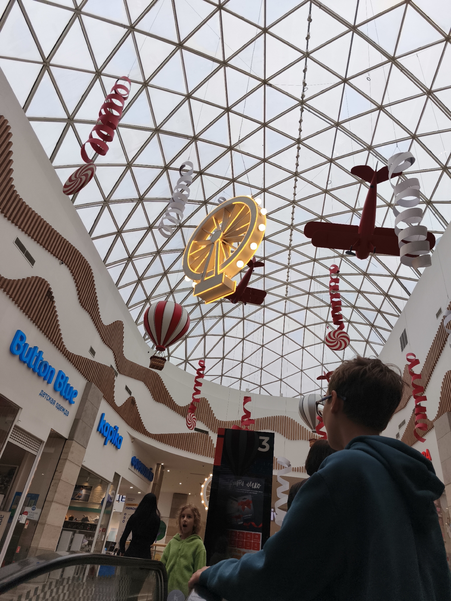 The image depicts the interior of a shopping mall with a geodesic glass roof. Suspended from the ceiling are decorative elements, including red and white spiral ribbons, a red biplane, a Ferris wheel illuminated with lights, and a hot air balloon. Storefronts with prominent signage like "Button Blue" and "kopika" are visible along the walls. Two individuals are partially visible in the foreground, with one wearing a blue hoodie and the other a green hoodie. A third person in black is also visible on an escalator. The scene appears to be captured from an upward-angle perspective, focusing on the ceiling decorations and the mall's architecture.
