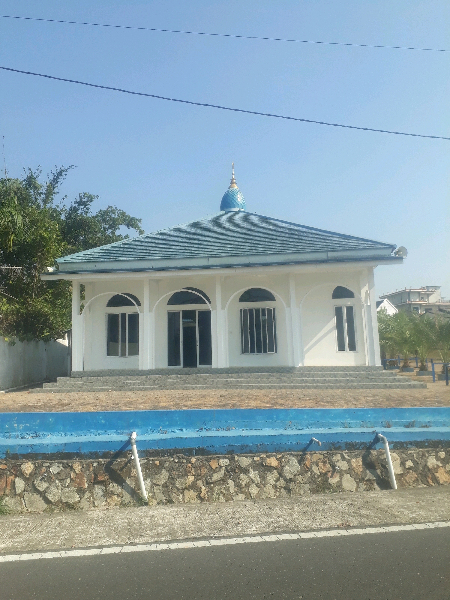 The image shows a white building with a blue dome and a blue-tiled roof, likely a mosque, situated in Pangkalpinang, Indonesia. The building features arched windows and a set of stairs leading to its entrance. In front of the building, there is a blue wall and a stone embankment. The area is surrounded by greenery, including bamboo trees on the left and palm trees on the right. A road runs in front of the embankment. The sky is clear and blue, indicating a sunny day. The image does not show any people.