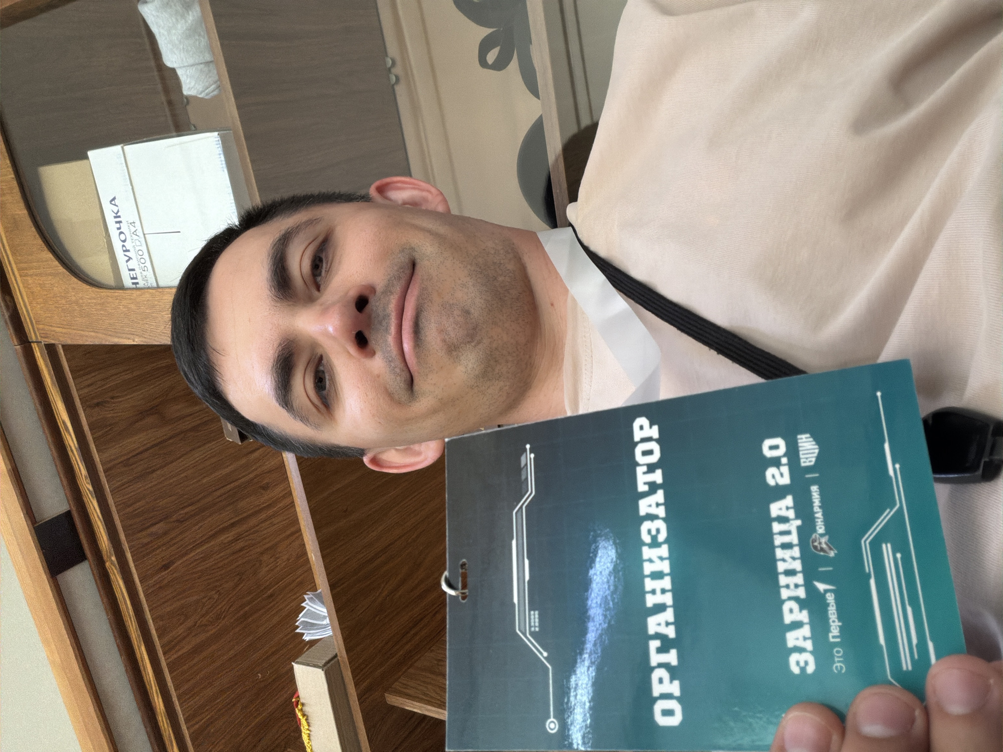 A man is holding a teal-colored badge that reads "ОРГАНИЗАТОР ЗАРНИЦА 2.0". The badge also has text that reads "Юнармия | ВДИН". The man has short dark hair and a slight smile. He is wearing a light-colored shirt with a white collar. The badge is attached to a black lanyard. The background includes a wooden cabinet and wall. There is text on a box on the top shelf that reads "ЧЕГУРОЧКА". The location is Забегалово, Russia.
