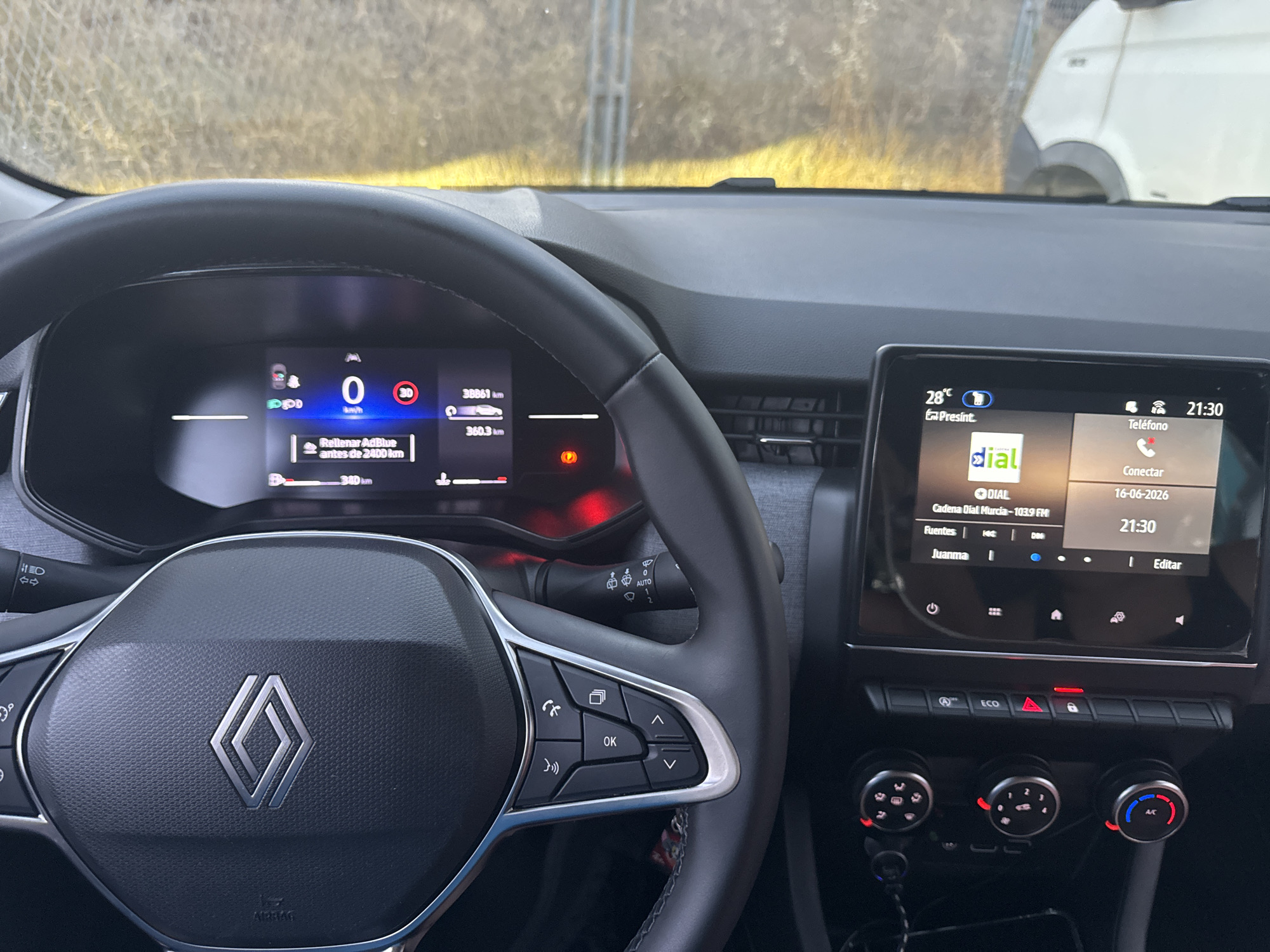 The image shows the interior of a car, likely a Renault, viewed from the driver's perspective. A digital dashboard displays vehicle information, including speed (0 km/h), distance traveled (38861 km), and a reminder to refill AdBlue. The steering wheel, with the Renault logo, is prominent in the foreground, featuring control buttons on the left side for calls and menu navigation. To the right, a central touchscreen display shows the current time (21:30), temperature (28°C), and radio information, including the station "Cadena Dial Murcia-103.9 FM" and the date "16-06-2026". Below the touchscreen are climate control knobs and buttons, including an "ECO" mode button. The background visible through the car's windows is an outdoor scene with dry grass and a fence, suggesting a location outside of a dense urban environment. The mention of "Murcia" in the radio station name strongly indicates the car is in or near the city of Murcia, Spain.