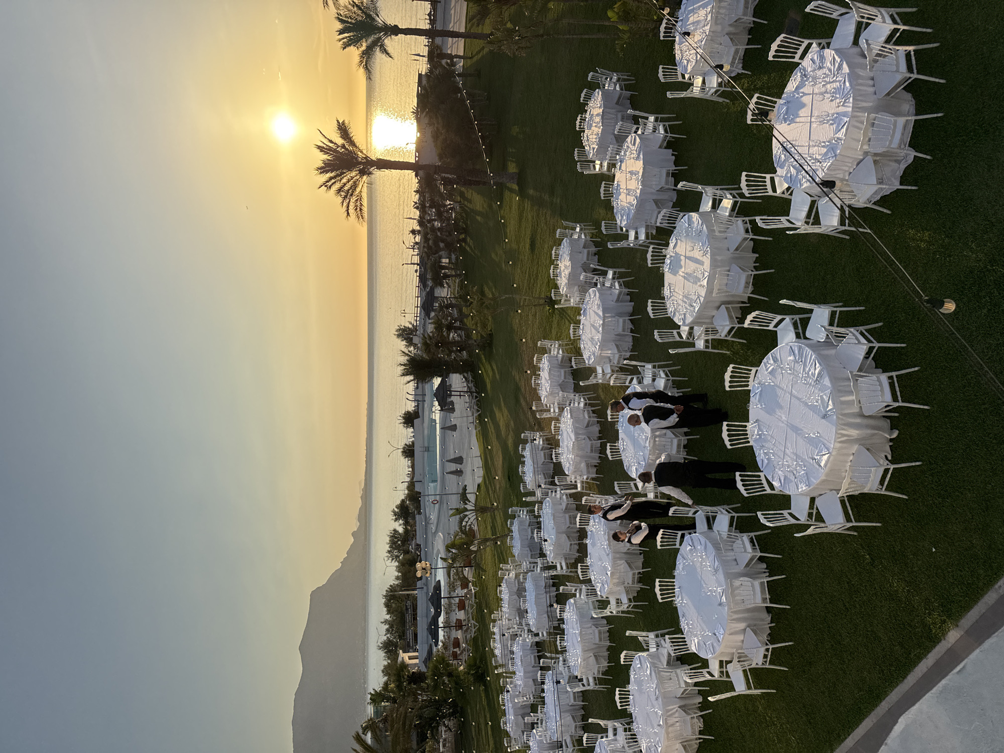 This image depicts an outdoor event setup at a coastal venue in Isola delle Femmine, Italy, during sunset. The low sun is a bright, intense golden orb on the horizon, casting a strong reflection across the calm sea. The sky transitions from a light blue overhead to warm yellow and orange hues near the horizon. A prominent, dark-silhouetted mountain range is visible in the far distance on the left.
Closer to the shore, a sandy beach area is apparent, alongside a large, irregularly shaped swimming pool surrounded by sun loungers and dark umbrellas. A sign indicating "Pizzeria Ristorante Hamadan Beach" is visible near the pool. Several palm trees are interspersed throughout the landscape between the pool area and the sea.
The foreground and mid-ground are covered by a well-maintained green lawn where numerous tables are arranged for an event. These tables are a mix of round and oval shapes, uniformly draped with white tablecloths and paired with white chairs. Each table setting includes multiple glasses and white napkins. String lights with exposed bulbs are strung across the grassy area above the tables.
Several male staff members, uniformly dressed in black trousers, white shirts, and black vests, are present among the tables. They are standing in various groupings, appearing to be engaged in coordination or final preparations for the event.