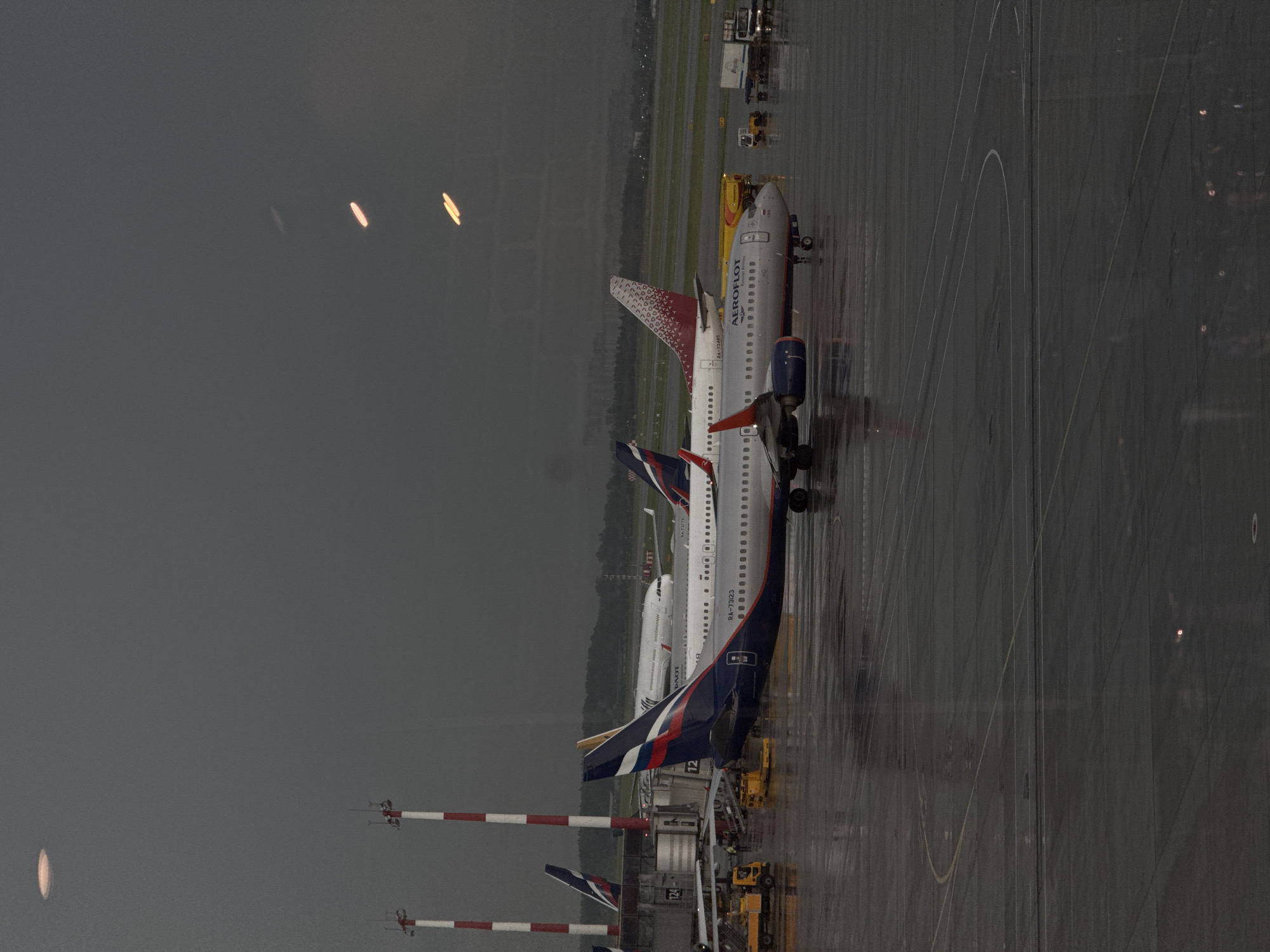 This is an outdoor, medium shot photograph taken at Sheremetyevo International Airport (SVO) in Khimki, Russia, likely on a cloudy or rainy day. The image features several airplanes parked on a wet tarmac. In the foreground, an Aeroflot Boeing 737-800 aircraft is visible, with its registration number RA-73123 partially obscured. Another Aeroflot airplane with a red and white patterned tail fin, identified by the registration RA-73775, is parked behind it. Further in the background, an Aeroflot Airbus A320 or A321 aircraft is also present. The tarmac shows reflections of the aircraft and the sky due to the wet conditions. In the distance, airport infrastructure such as jet bridges, service vehicles, and other parked aircraft can be seen. The sky is overcast, creating a somber atmosphere.