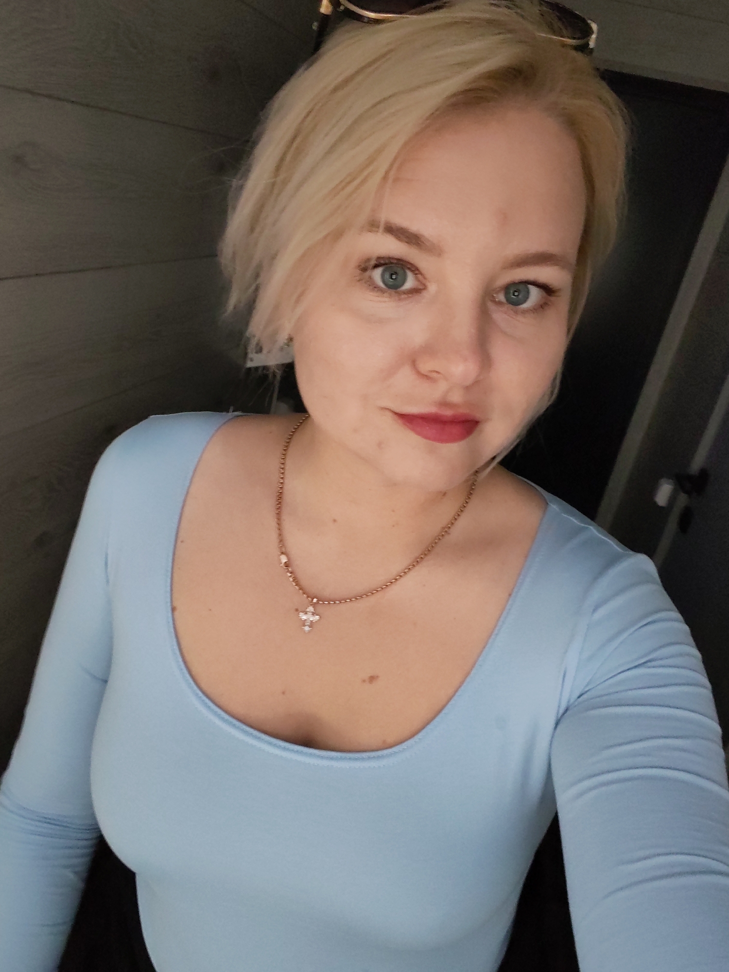 A blonde woman with blue eyes and light pink lipstick is captured in a close-up, chest-up indoor shot, looking directly at the camera. She has short, light blonde hair. Sunglasses with dark lenses and light frames are perched on top of her head. She wears a light blue, long-sleeved, scoop-neck top. A gold-toned chain necklace with a small, white-jeweled cross pendant hangs on her chest. The background on the left is a dark, horizontally paneled wall, while the right side shows a dark, featureless wall or door with a faint dark handle visible.