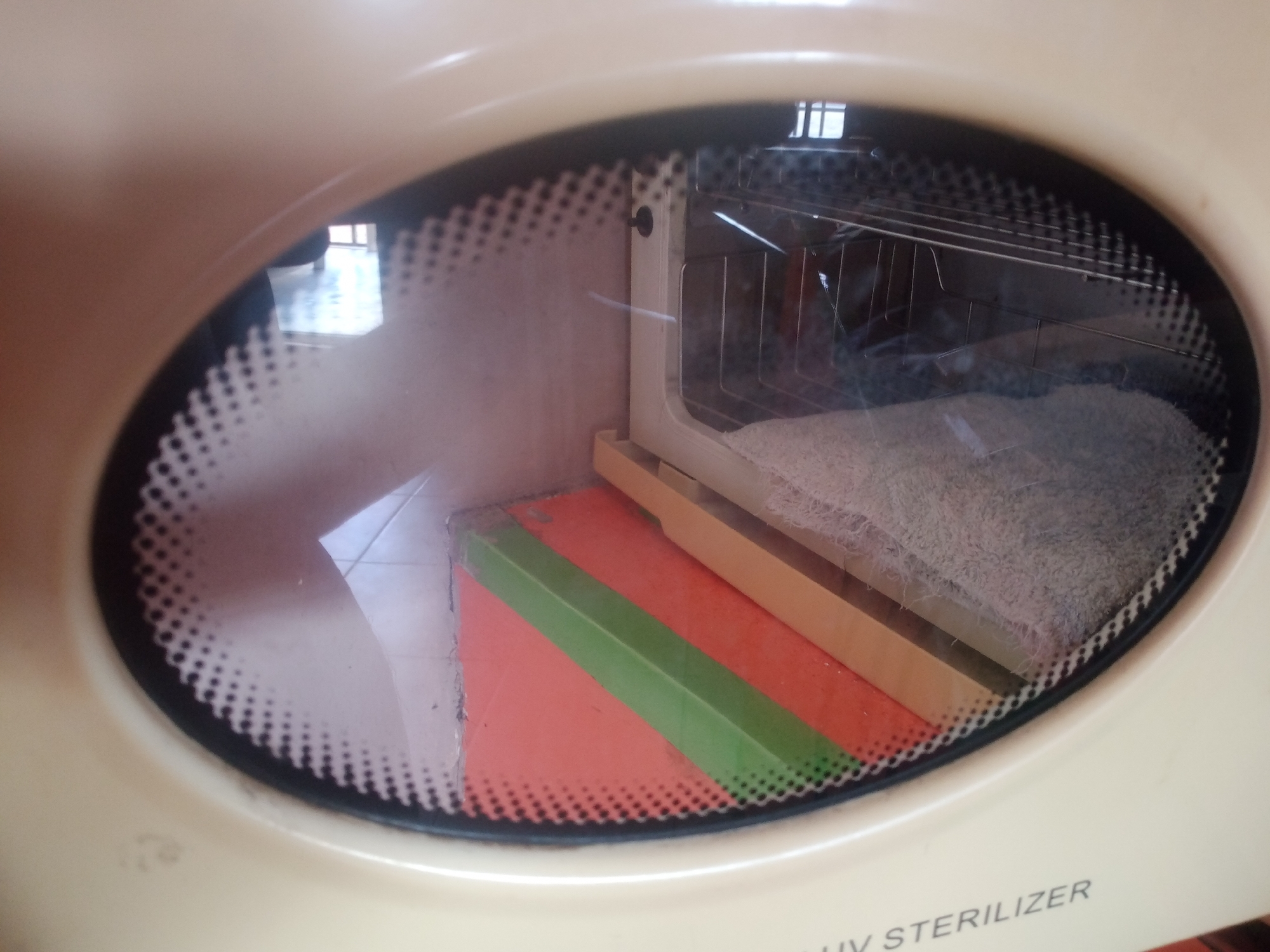 A tan-colored UV sterilizer has its door open, revealing the interior. Inside, there is a wire rack, a beige tray, and a white cloth. The sterilizer is placed on a surface that appears to be a floor or a table, with a glimpse of a window in the background. The lighting suggests it is daytime. There are no people or animals visible in the image. The sterilizer is likely located in a domestic or commercial setting in Jalingo, Nigeria.