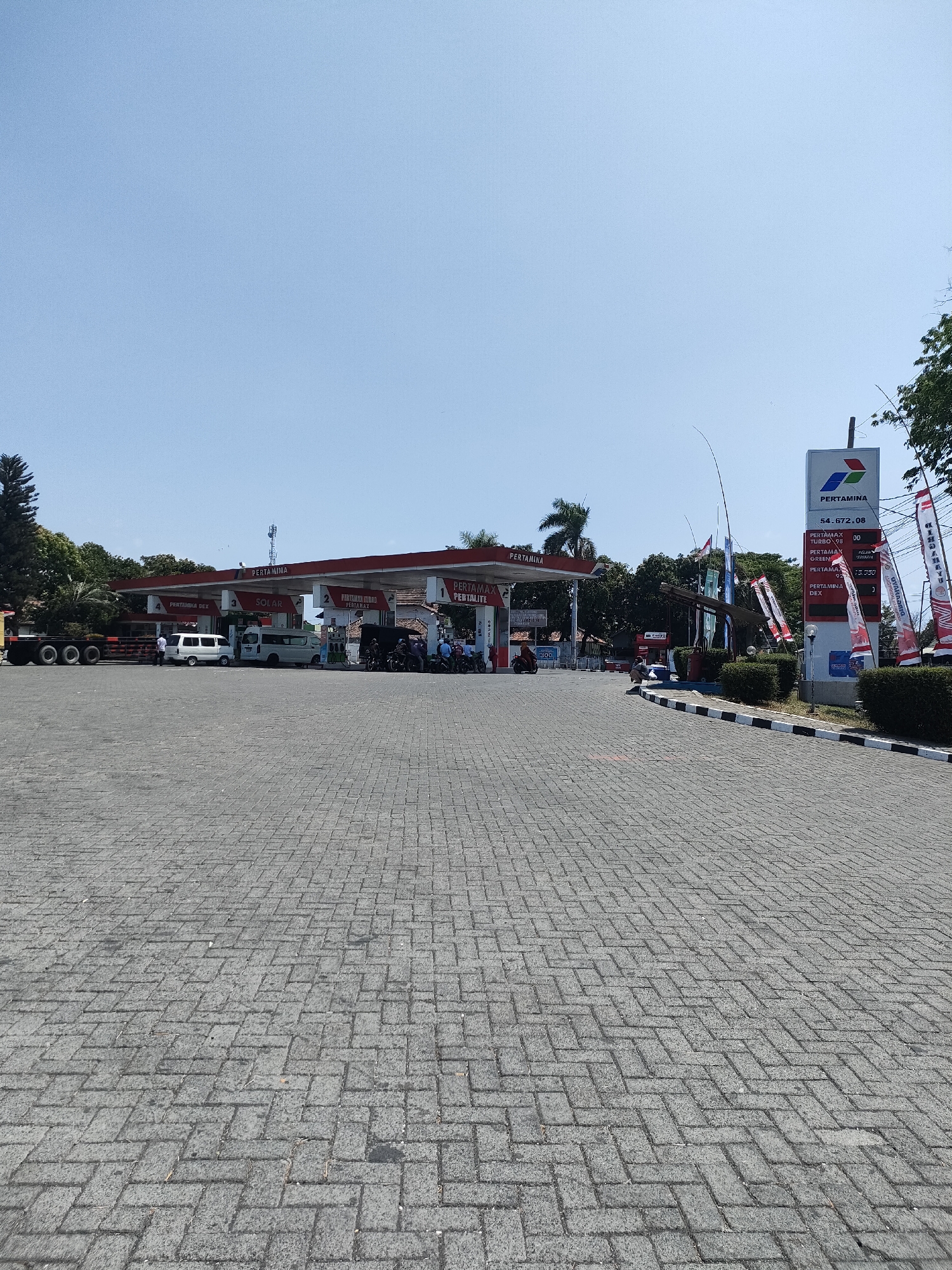 The image captures a wide view of a Pertamina gas station in Curahdringu, Indonesia, under a clear blue sky. The foreground is dominated by a grey paver stone parking lot. A Pertamina gas station canopy with red trim and white support columns is visible in the mid-ground, with several fuel pumps beneath it. A tall sign displaying "PERTAMINA" and fuel prices stands to the right of the canopy. Various vehicles are present, including a large truck with multiple axles, several vans, and motorcycles. Some people are seen standing near the pumps and vehicles. Flags with "PERTAMINA" branding are displayed prominently. The environment appears to be a typical roadside setting with some trees and vegetation visible in the background.