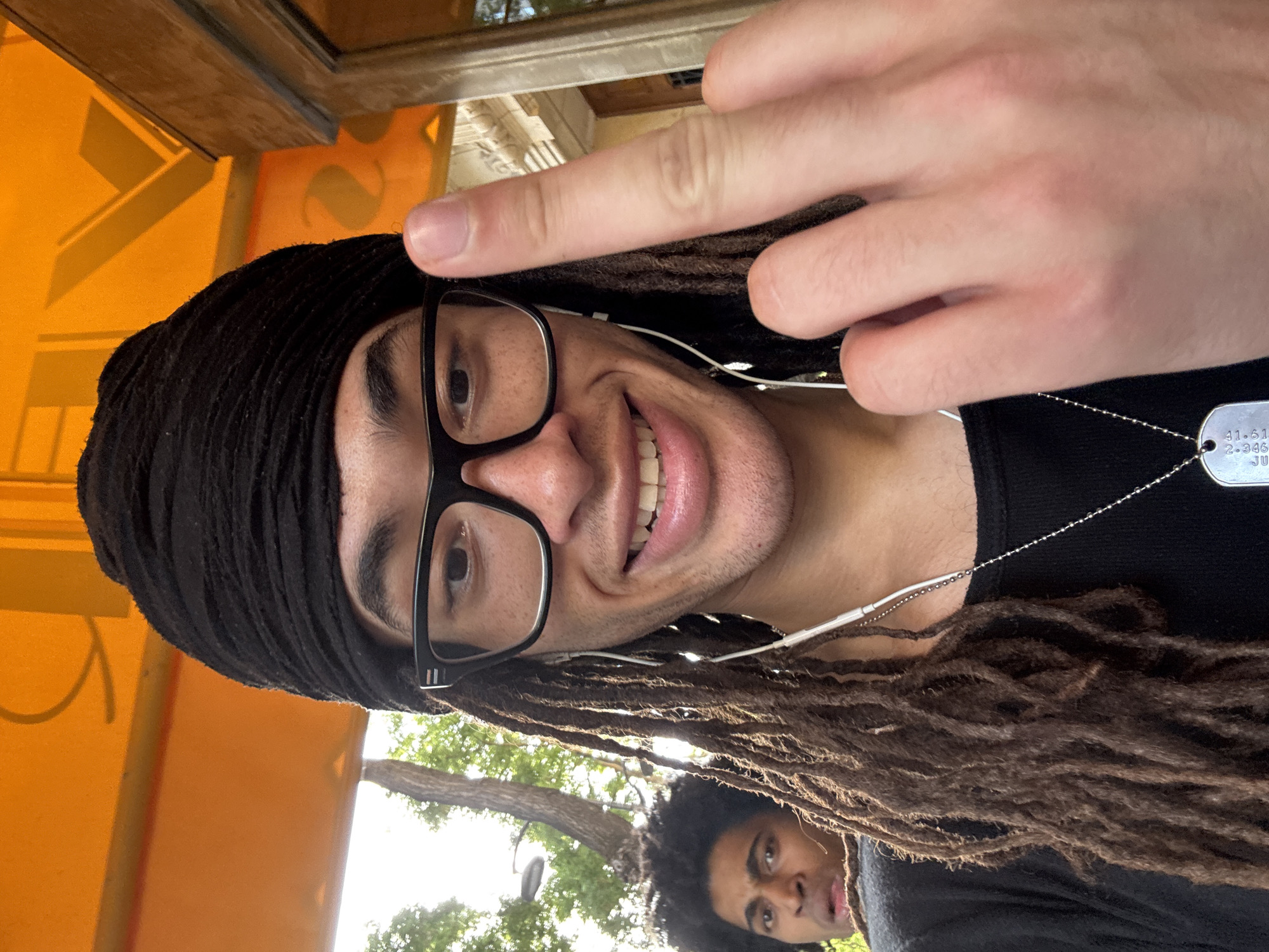 A selfie-style photograph features a person with dreadlocks and glasses, wearing a black head covering and a black shirt. A silver dog tag necklace is visible. The person is smiling and holding up their hand near their face. A second person with curly hair is partially visible at the bottom of the frame. The background includes an orange structure and glimpses of trees and sky, suggesting an outdoor location in Paris, France.