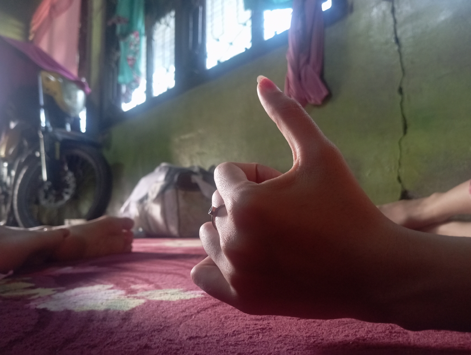 In Rejosari, Indonesia, a close-up shot focuses on a person's hand, which is held up with the thumb extended, suggesting a "thumbs up" gesture. A ring is visible on one of the fingers. The hand is positioned in the foreground on a textured red surface, possibly a rug or blanket. In the blurred background, a motorcycle is partially visible, along with some clothing hanging from what appears to be a window frame. A crack is noticeable on the light green wall. To the left, another person's feet are visible, also resting on the red surface. The overall environment suggests an indoor, possibly residential, setting.